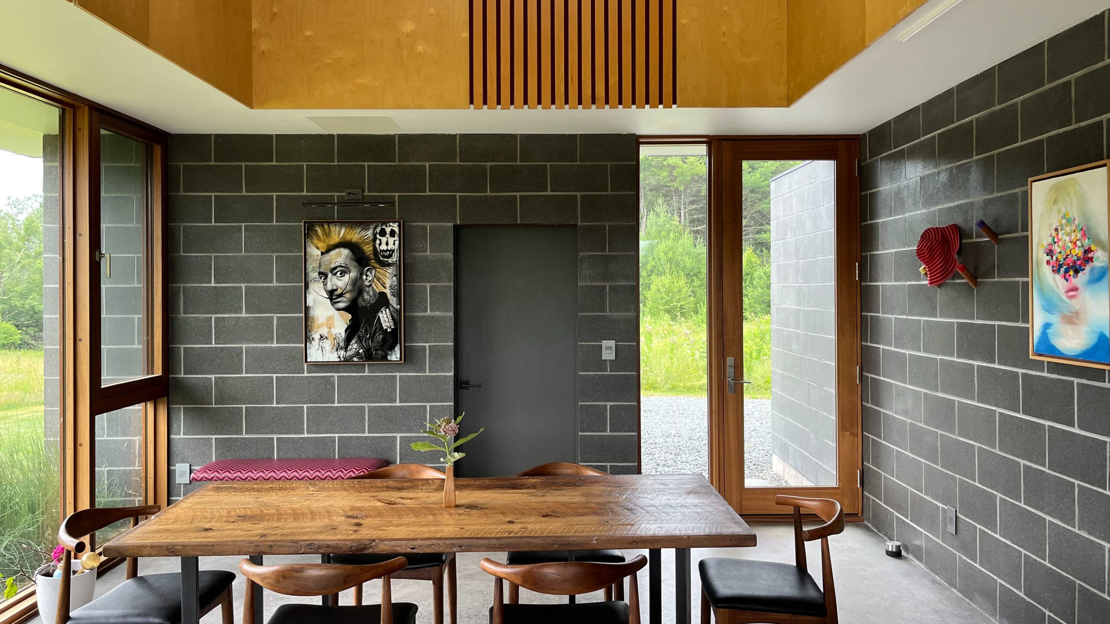 A modern dining area with a wooden table surrounded by dark chairs, a piece of wall art, and large windows overlooking a green landscape.