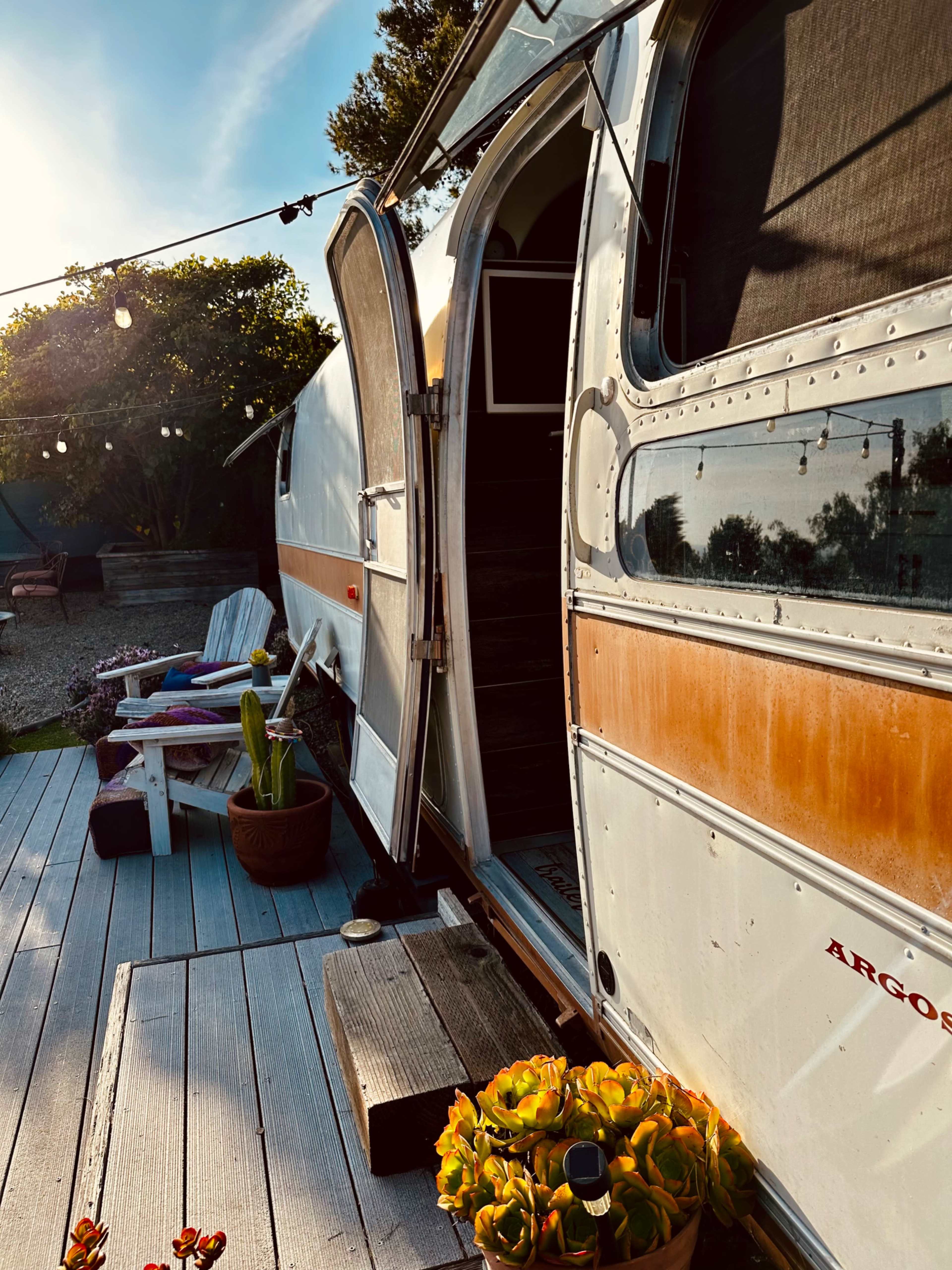 An Airstream trailer with an open door sits on a wooden deck adorned with potted plants and string lights.