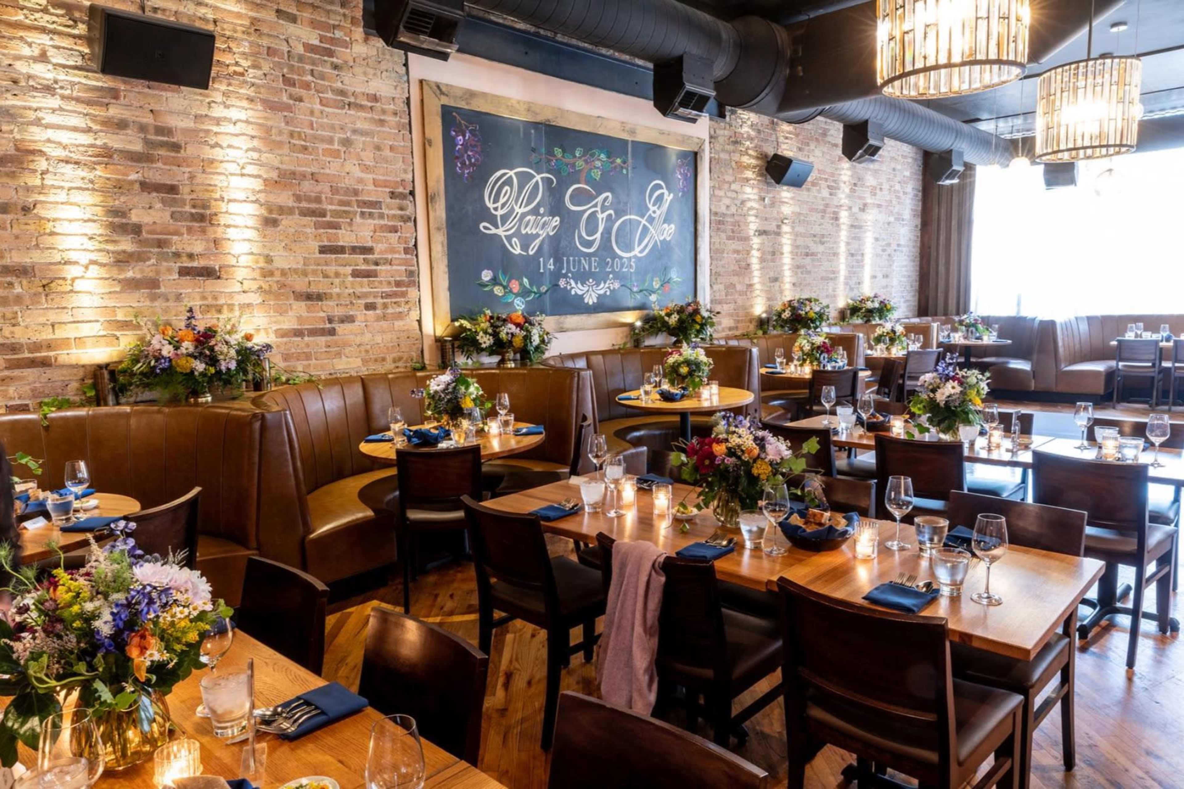 The image shows a warmly-lit restaurant interior featuring wooden tables arranged with floral centerpieces and blue napkins, alongside brown leather booths and a decorative chalkboard.