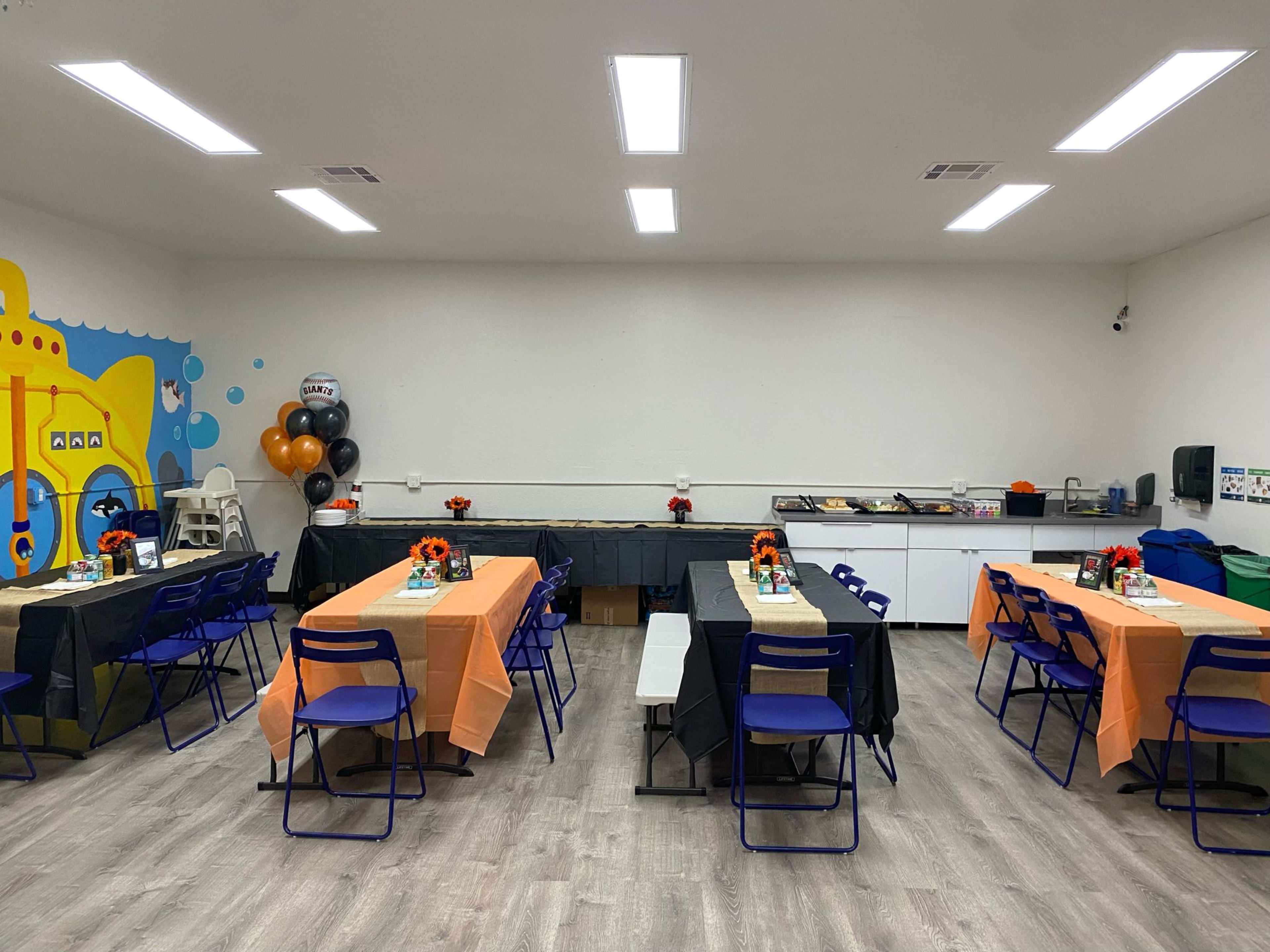 A brightly lit room with tables set for an event, featuring orange and black tablecloths and decorations, arranged alongside a wall with a colorful mural.