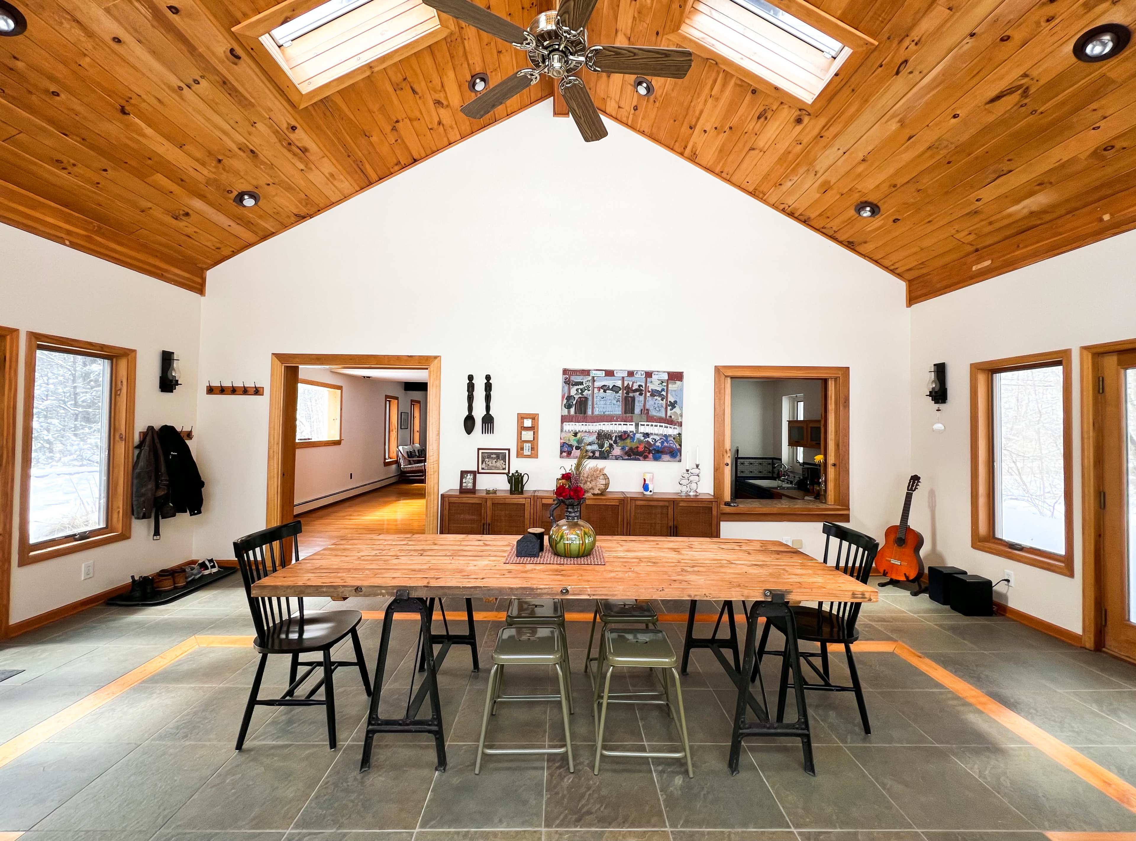 A spacious dining area features a long wooden table surrounded by black and silver chairs, with large windows and a wooden ceiling above.