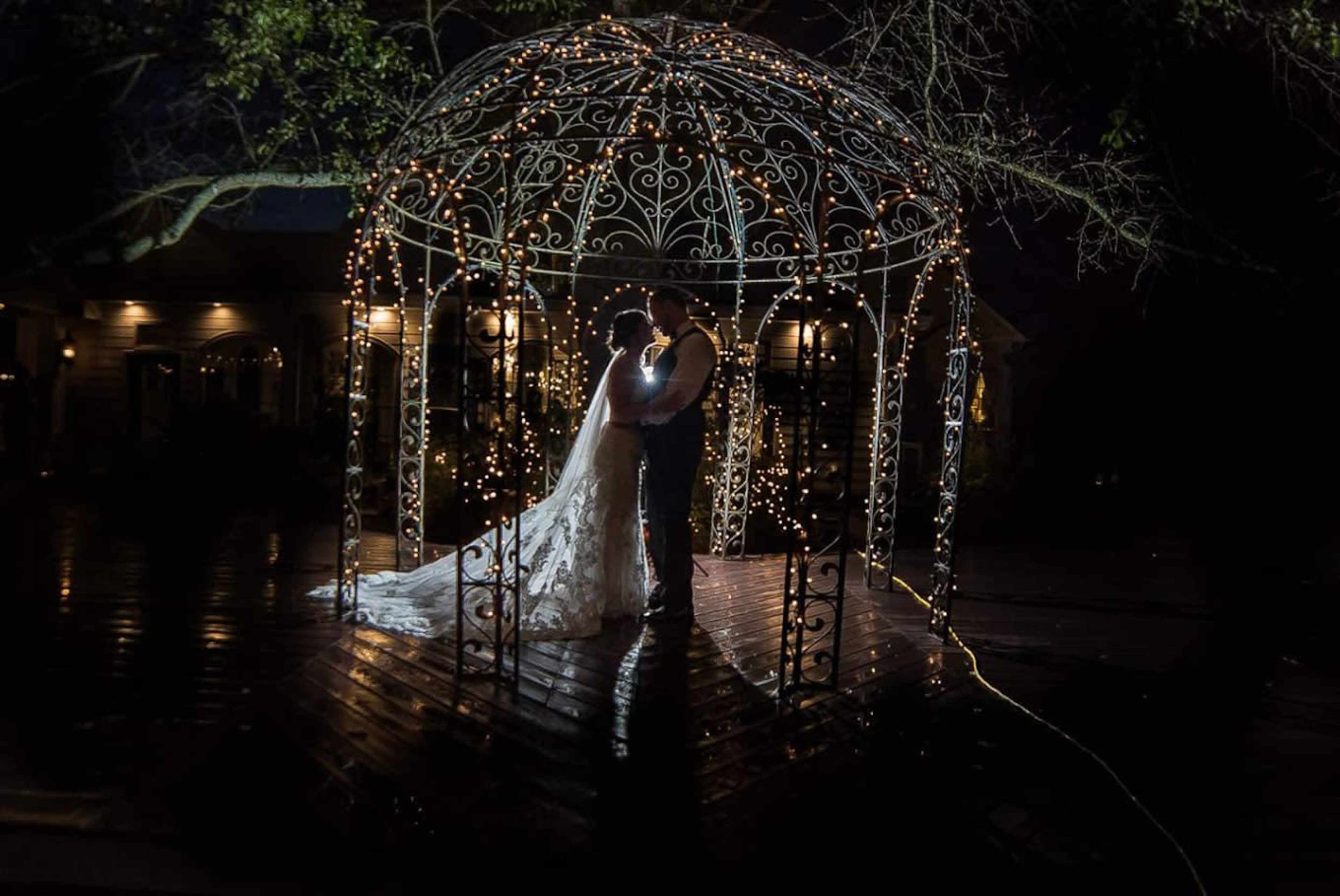 A couple stands under a lit gazebo at night, surrounded by a reflective wooden deck and trees.