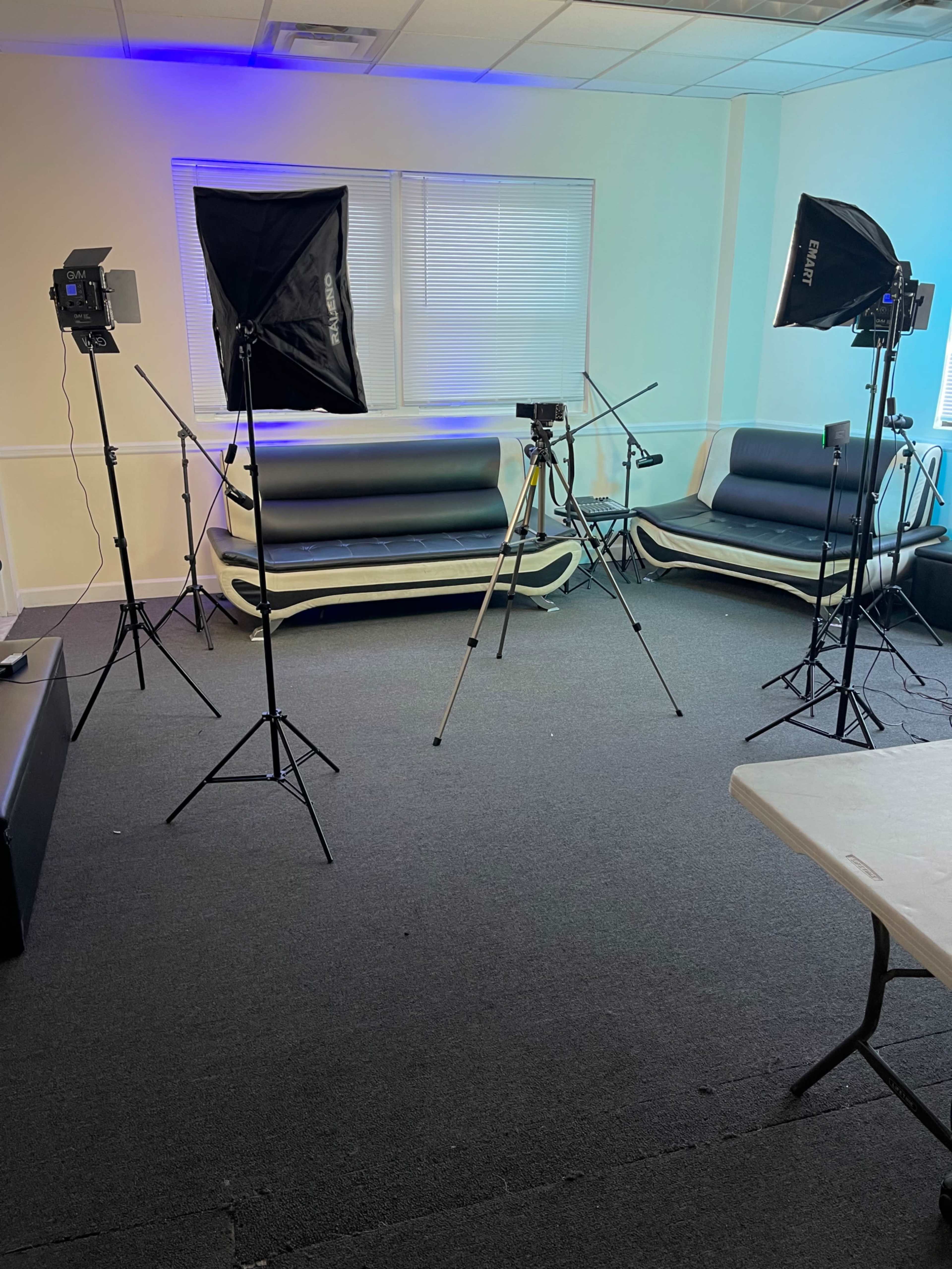 The image shows a room equipped with softbox lights and tripods positioned near two dark couches and a table.