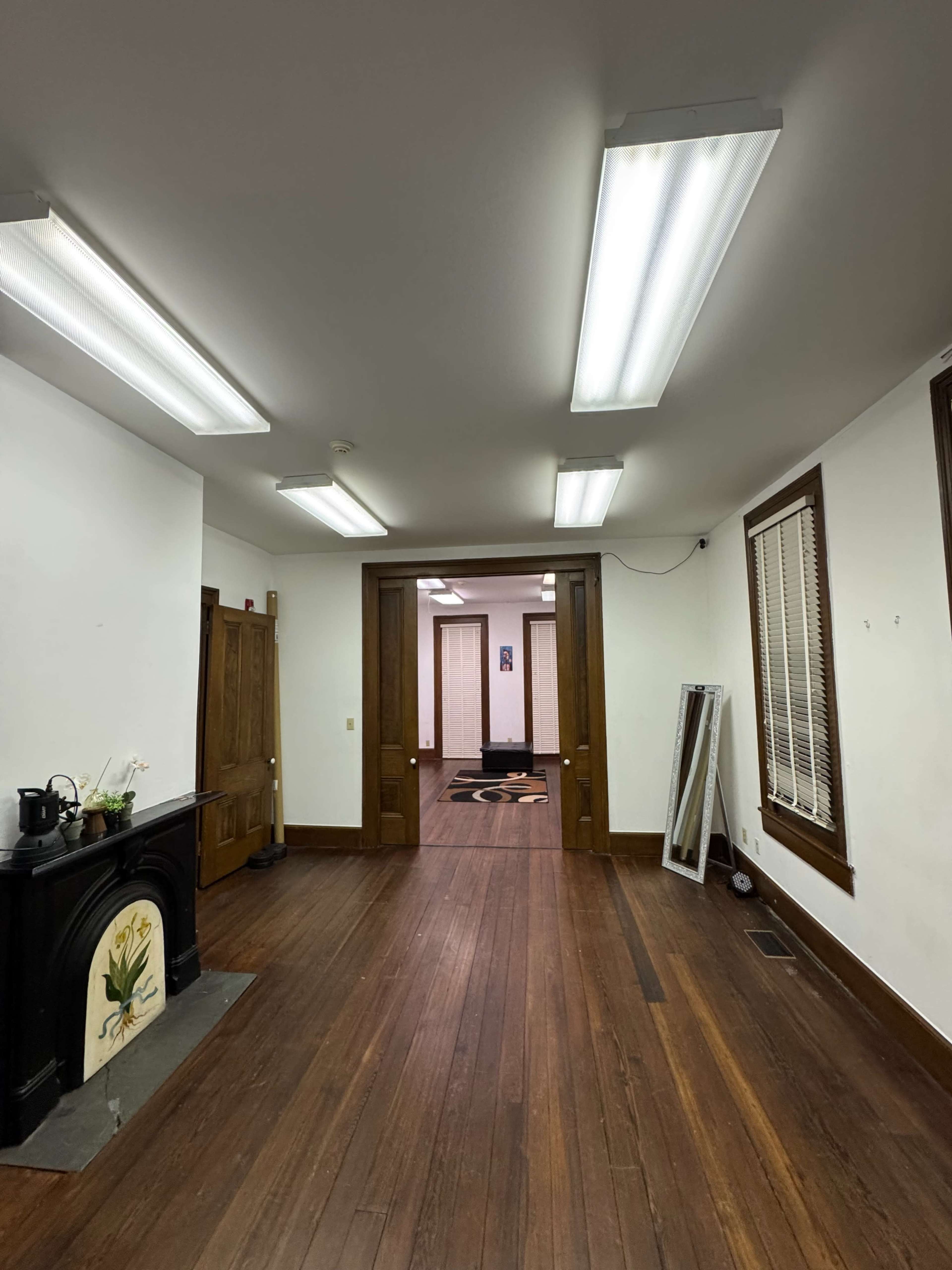 The image shows a spacious room with wooden floors, overhead fluorescent lighting, and a decorative fireplace on the left side.