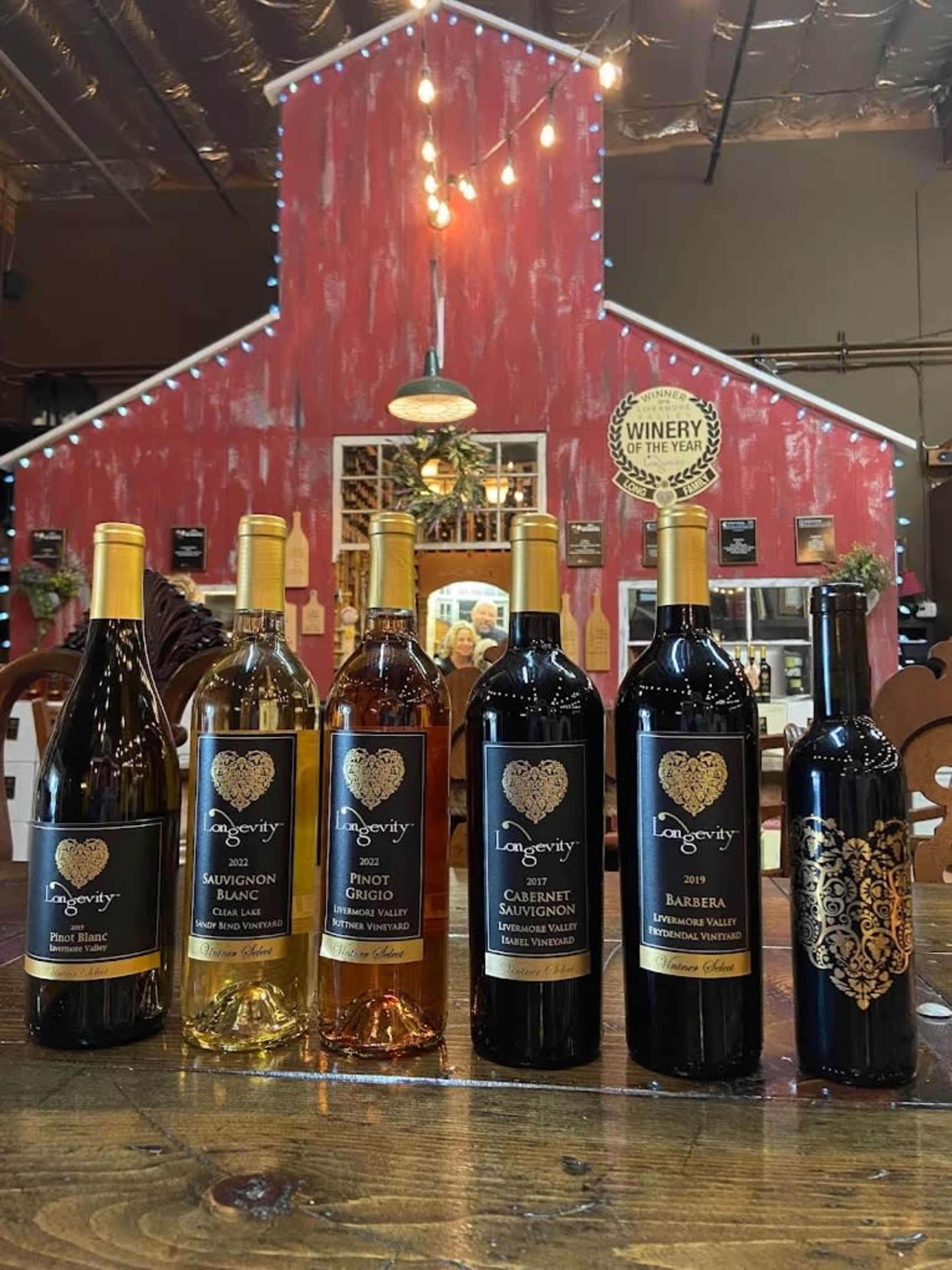 The image displays a collection of seven wine bottles arranged on a wooden table, with a rustic red barn backdrop and various images and shelves in the background.