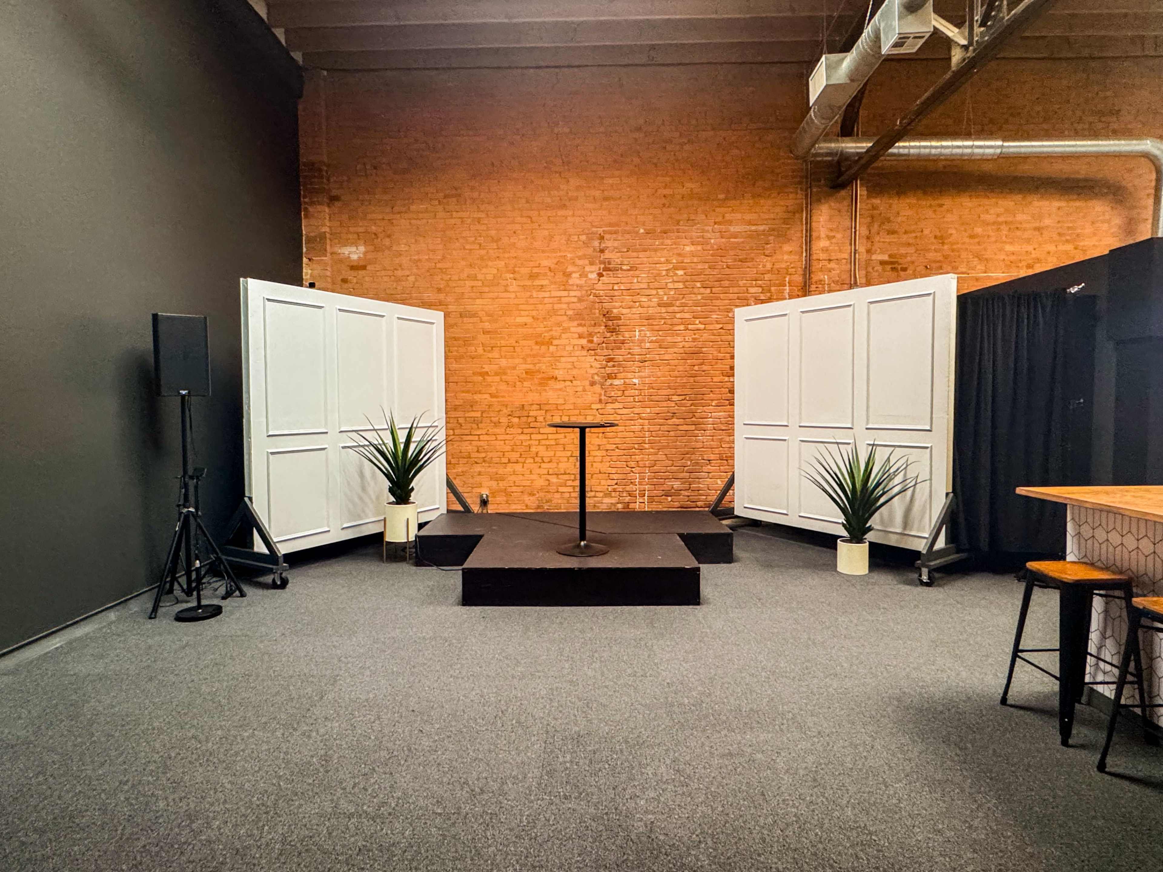 The image shows a small stage set up in a venue with a wooden platform, a single mic stand, potted plants on either side, and white folding doors against a brick wall.