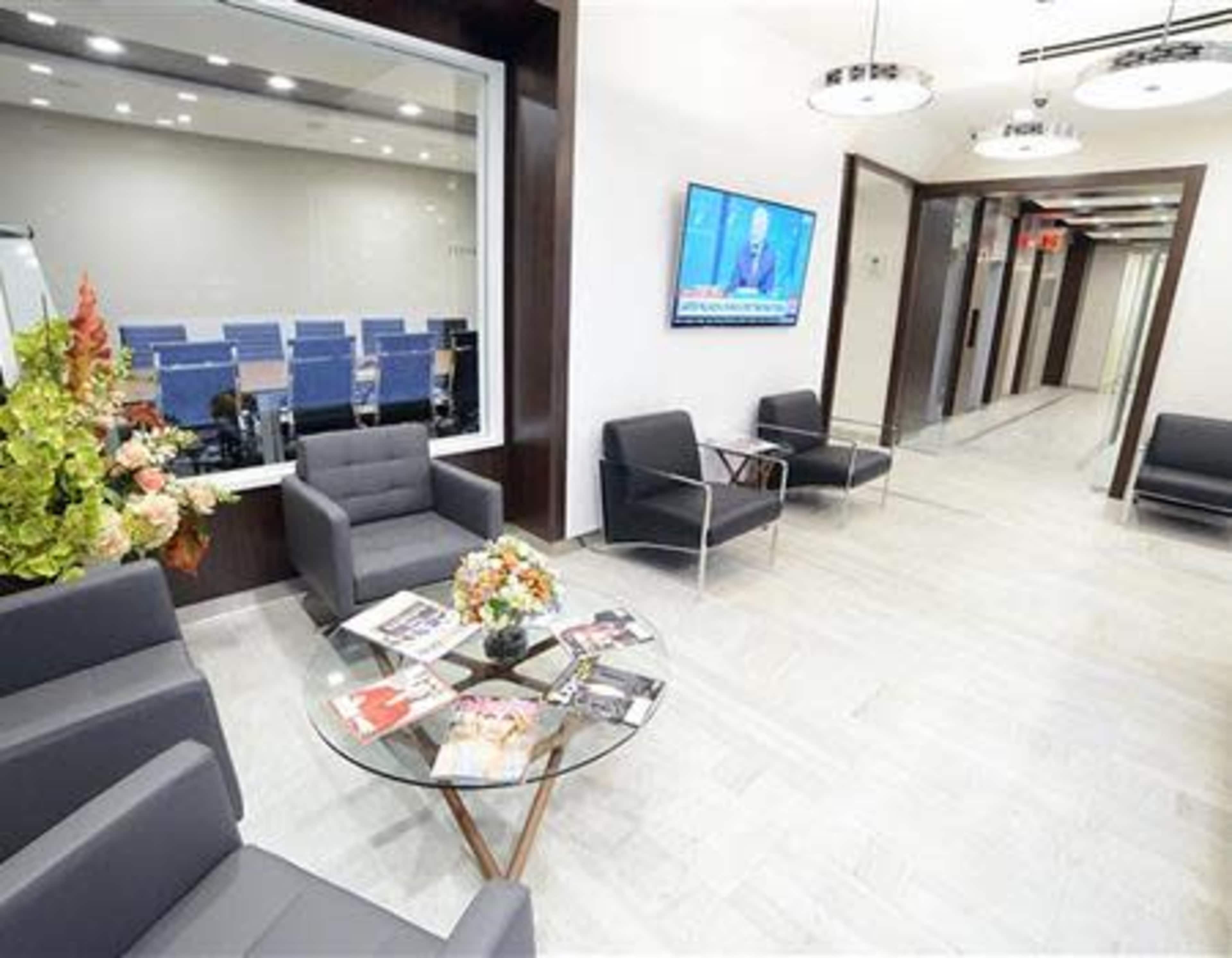 A modern office reception area with gray chairs, a glass table adorned with magazines, and a television displaying news, alongside a corridor leading to meeting rooms.