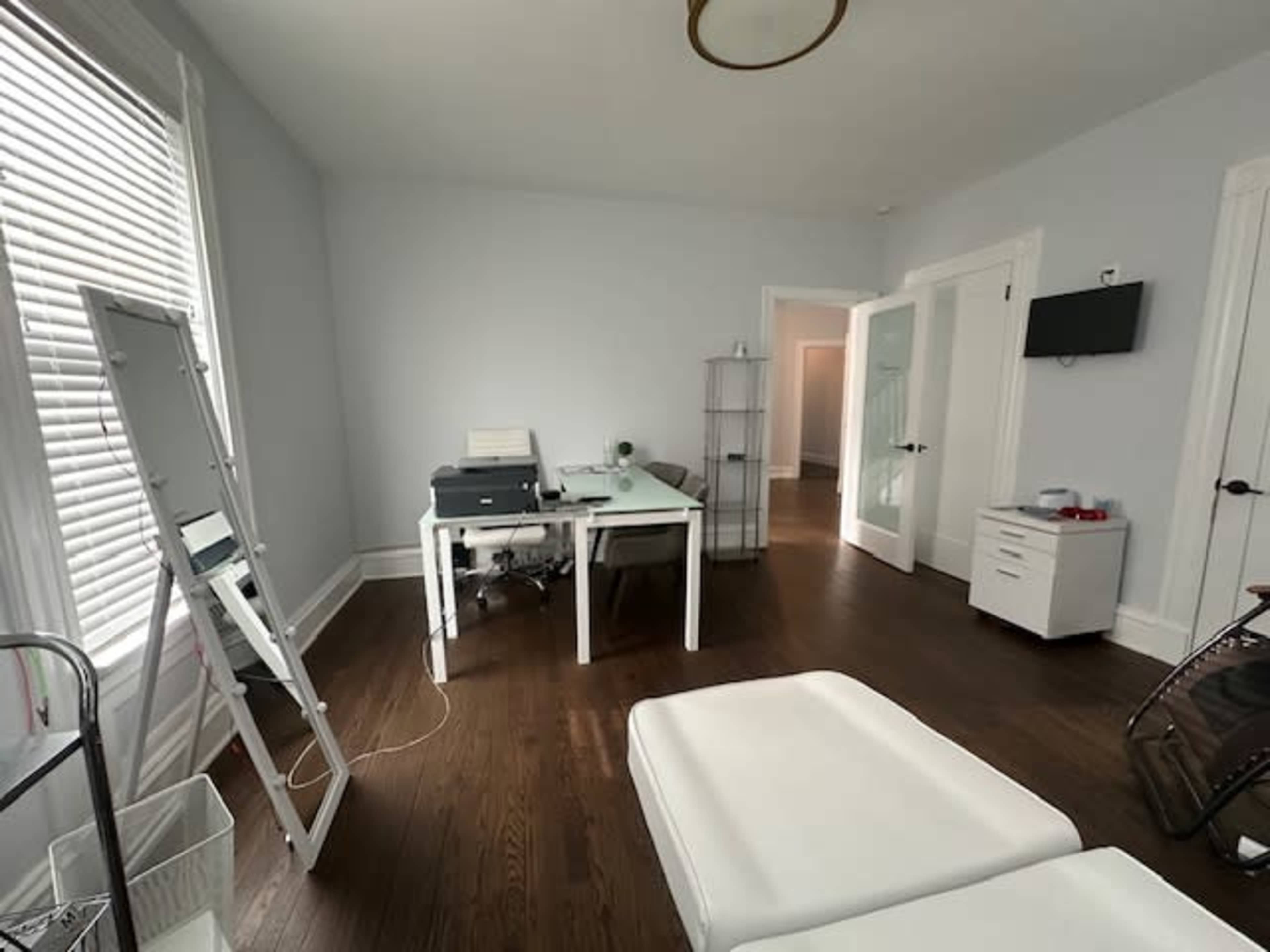 A well-lit room features a desk with a printer, a mirror, and two white treatment tables, along with a closet and a wall-mounted TV.