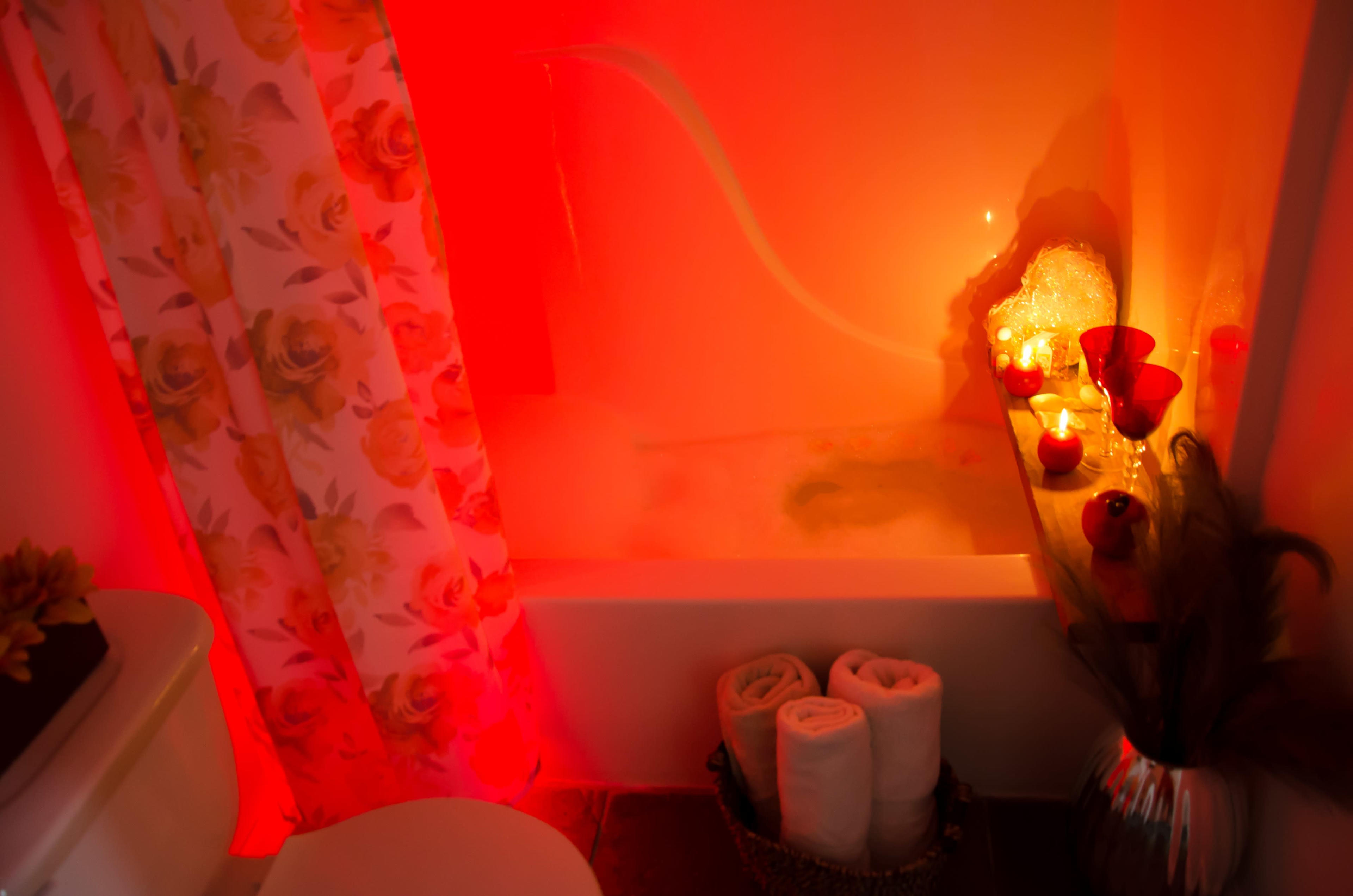 The bathroom features a bathtub illuminated by red lighting, surrounded by decorative towels and candles on a wooden shelf.