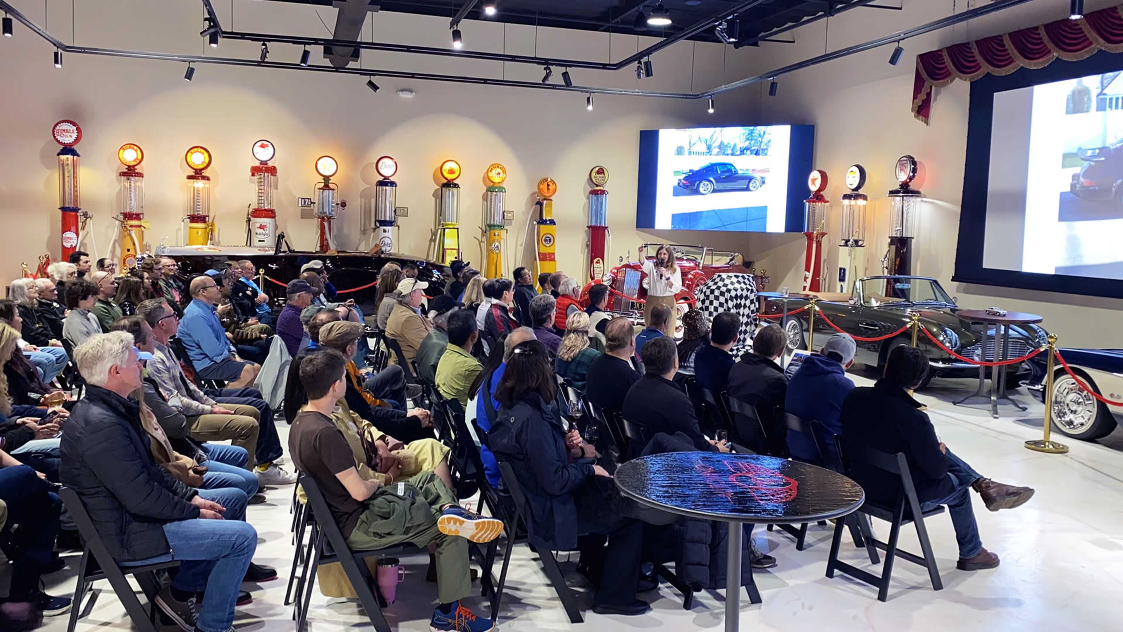 A large audience is seated in a showroom filled with vintage cars and gas station memorabilia, while a presenter speaks in front of a screen displaying images of cars.