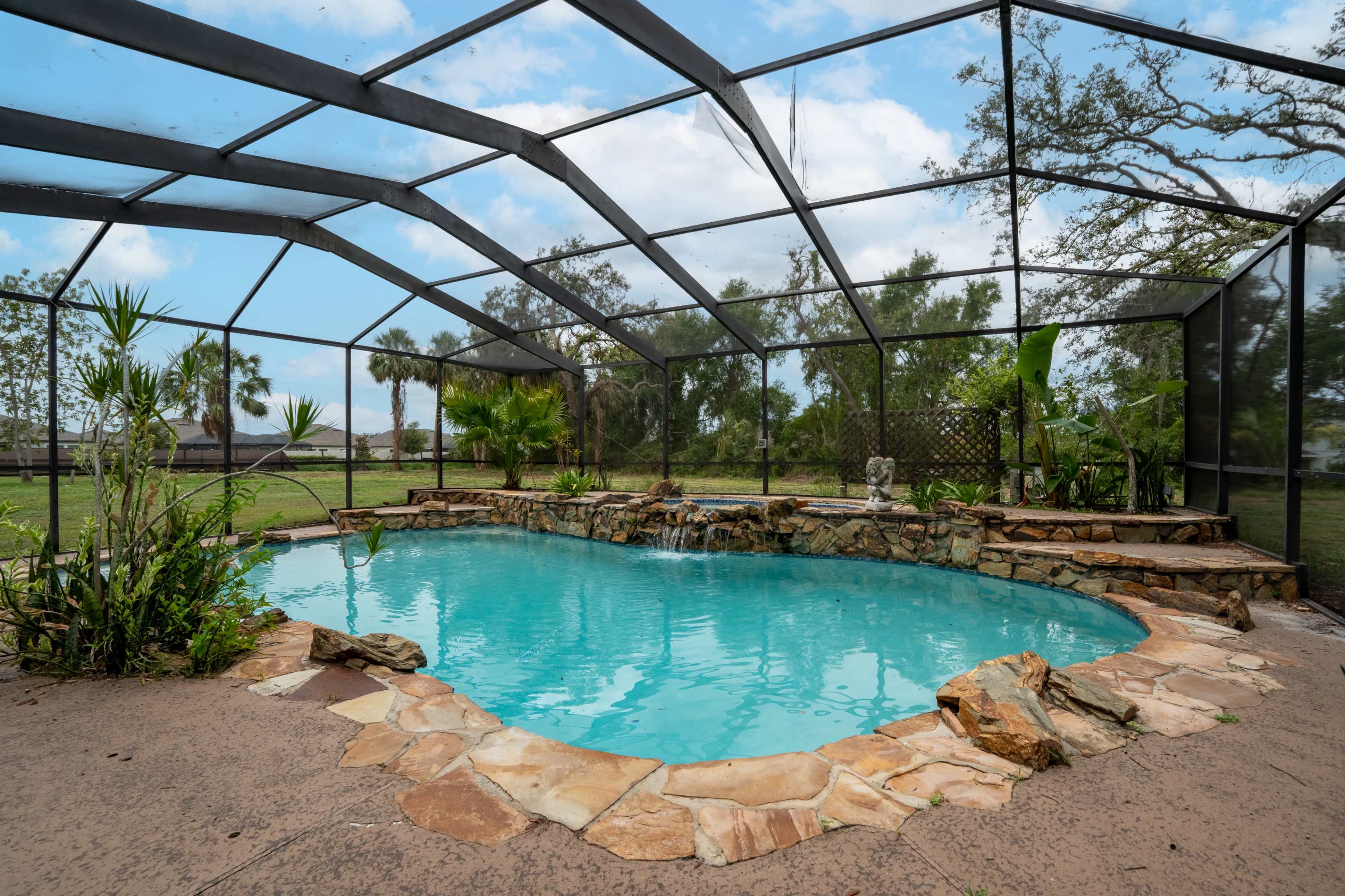 The image shows a rectangular swimming pool with a stone surround, enclosed by a mesh screen, and surrounded by greenery.