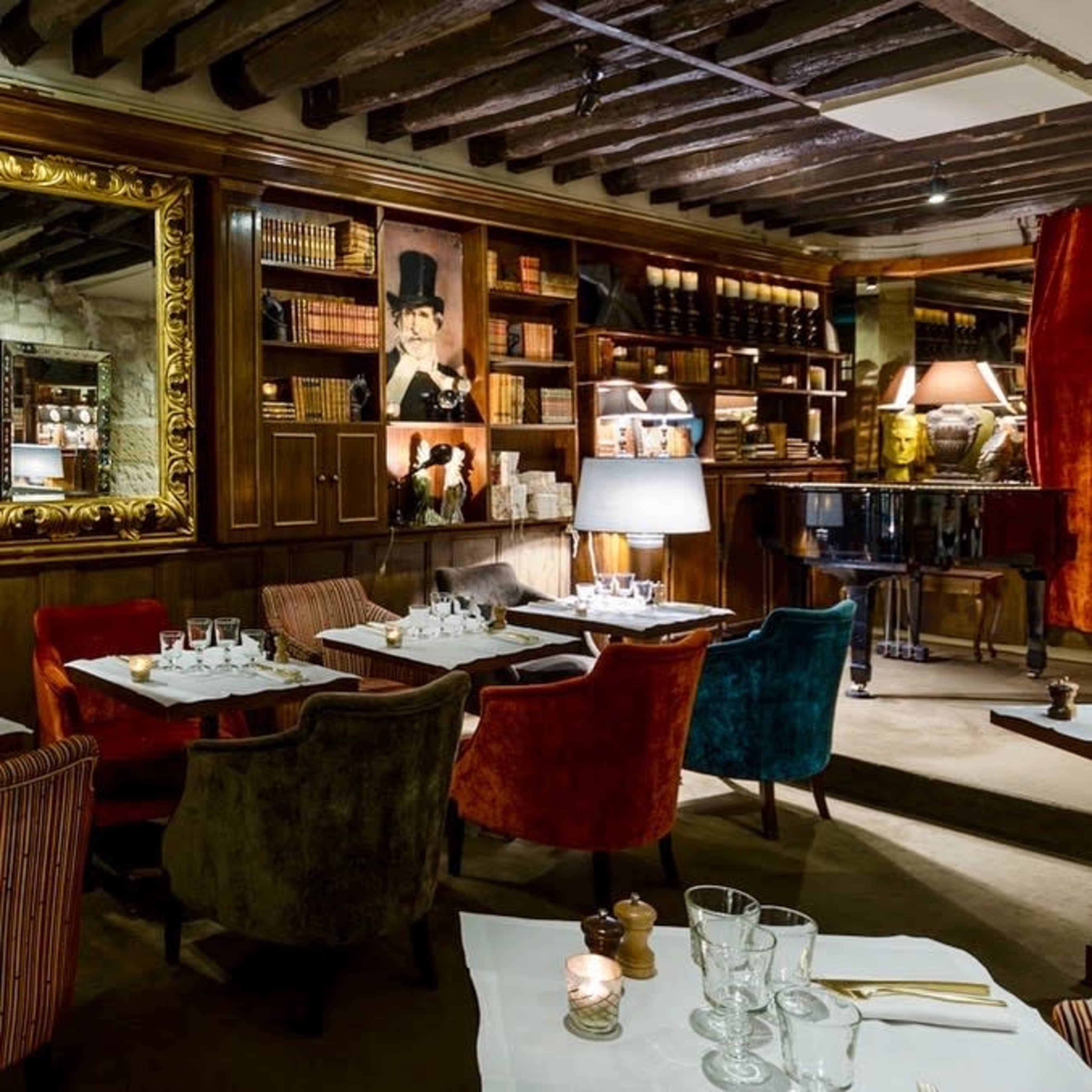 A cozy restaurant interior with wooden beams, plush chairs in various colors, a dining area with set tables, and a grand piano near a bookshelf filled with books and decorative items.