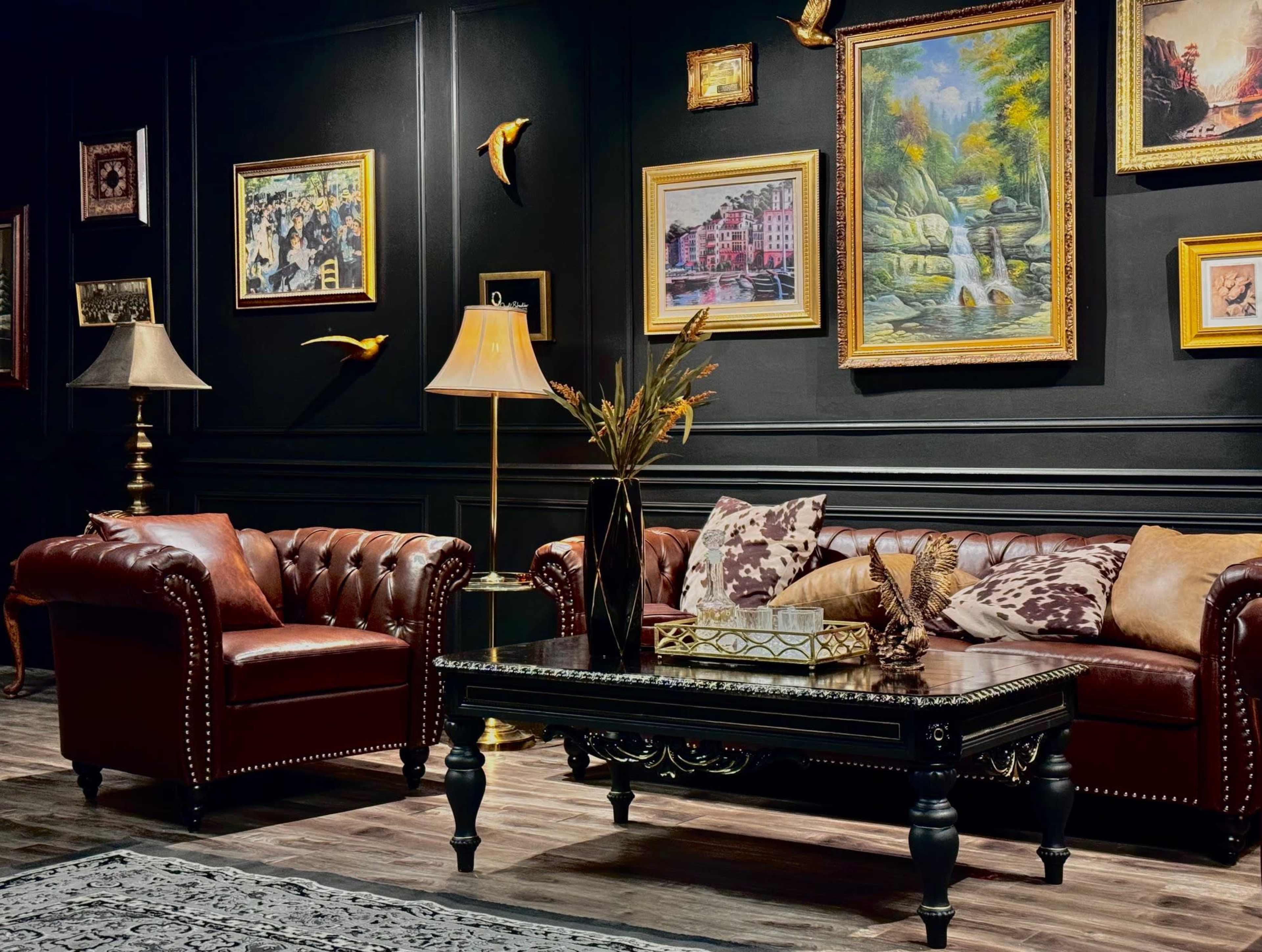 The image shows a living room with a dark wall adorned with various framed paintings, featuring a leather chesterfield sofa, an armchair, a decorative coffee table, and lamps.