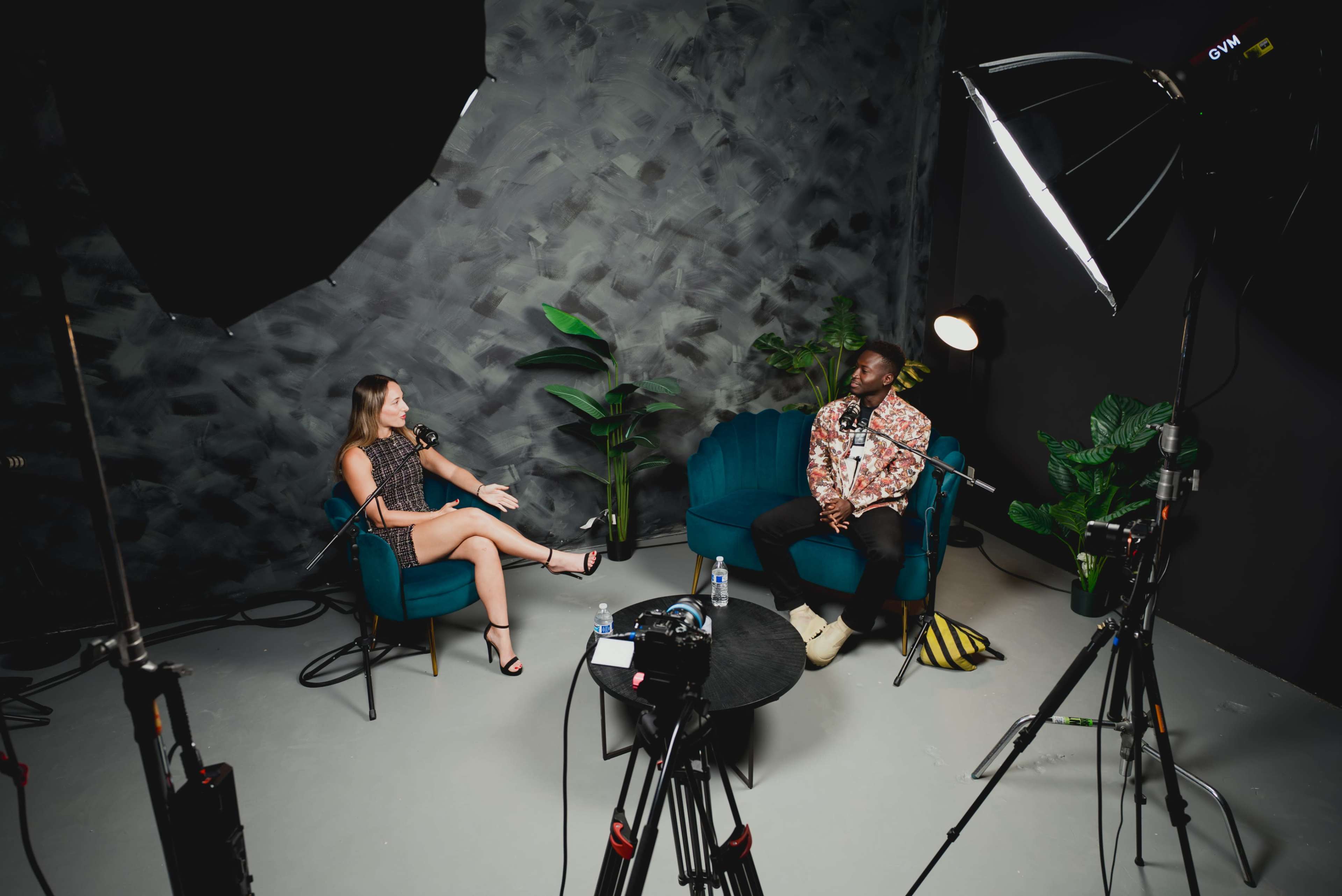 A woman and a man are seated in a recording studio, engaged in a conversation while surrounded by cameras and microphones.
