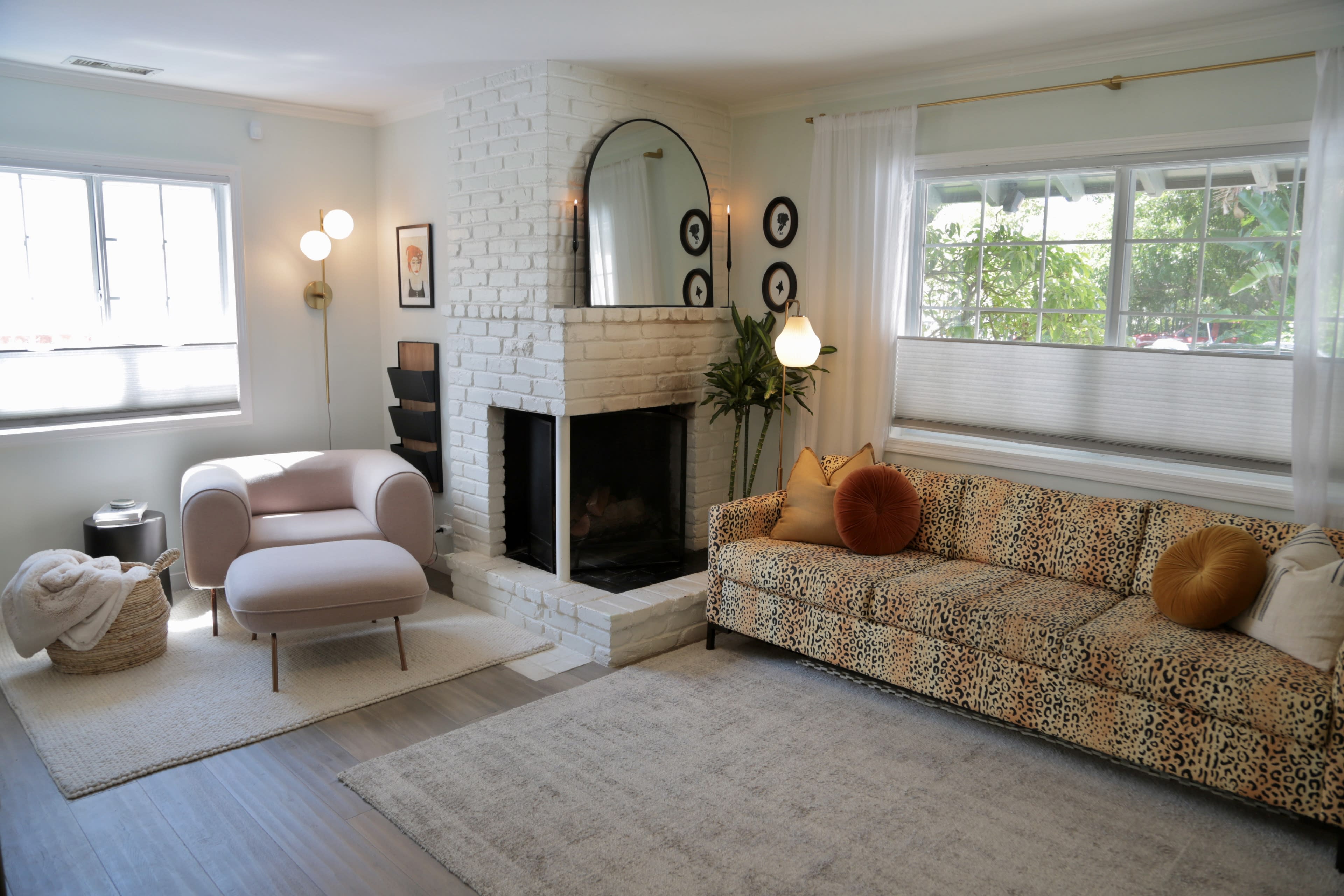 A living room featuring a white-brick fireplace, a beige animal-print sofa, a light pink armchair, and a large window with natural light.