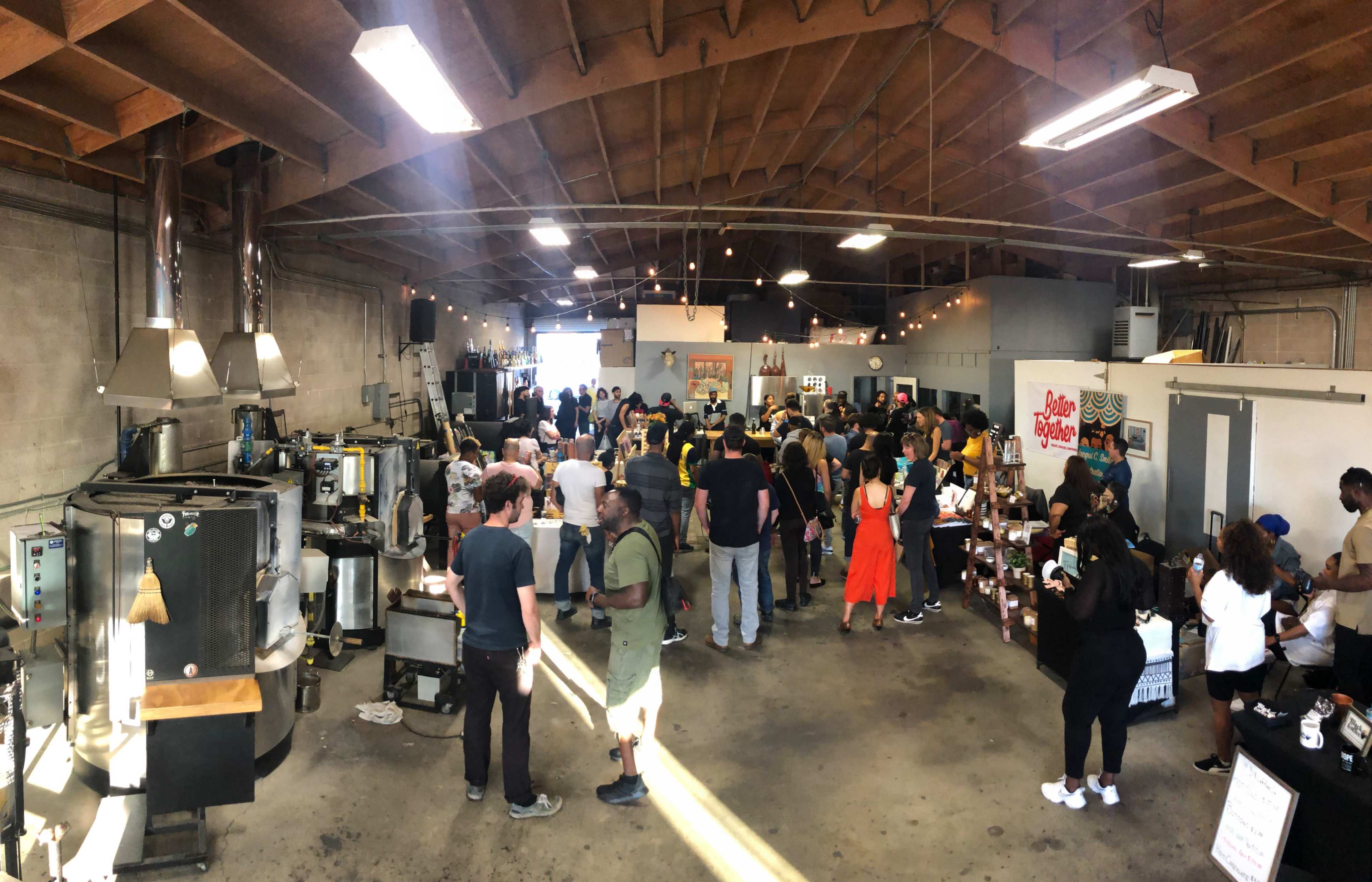 A large crowd gathers in a spacious coffee roastery, surrounded by roasting equipment and tables featuring various coffee products.