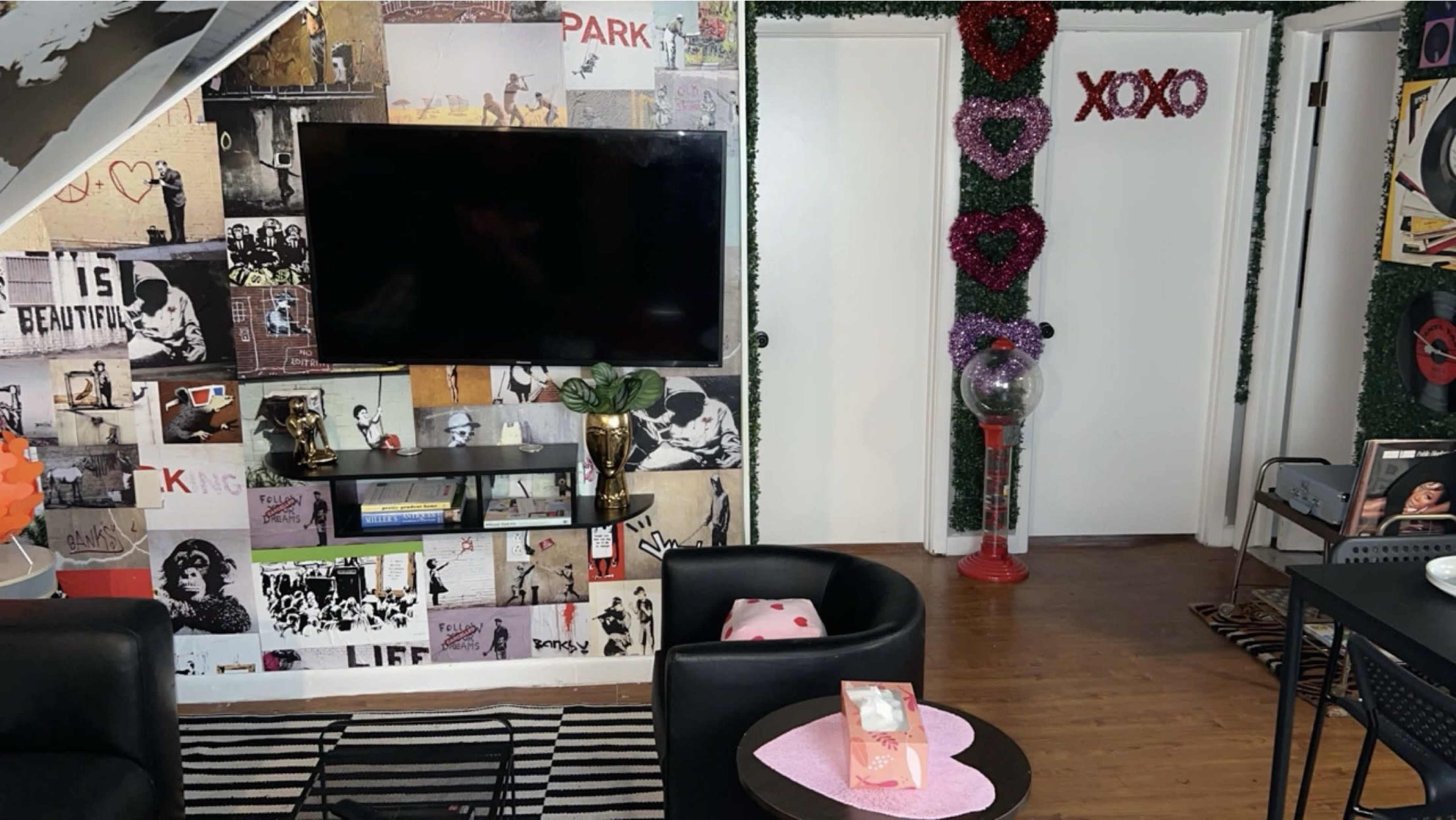A living room features a wall covered in a collage of posters, a wall-mounted TV, a coffee table, and a door adorned with heart decorations.