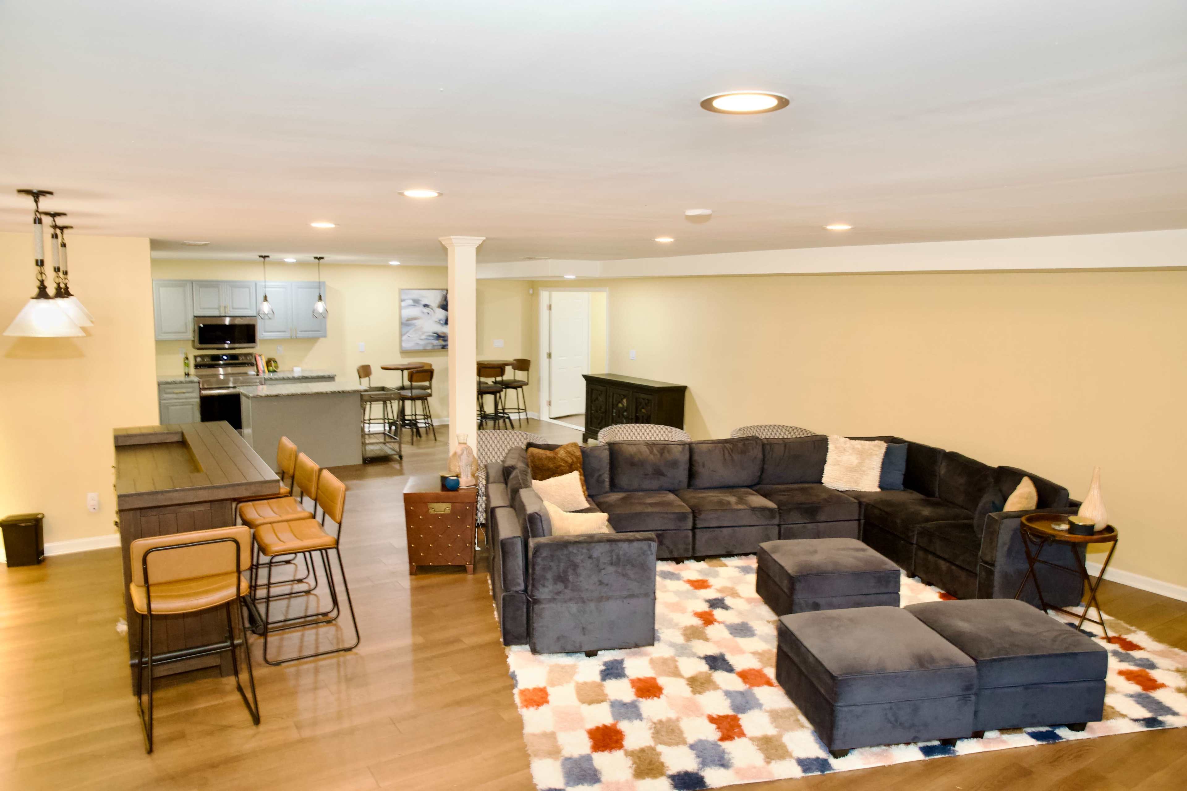 The image shows a spacious living area with a large sectional sofa, a coffee table, and a dining space with bar stools, adjacent to a kitchen.