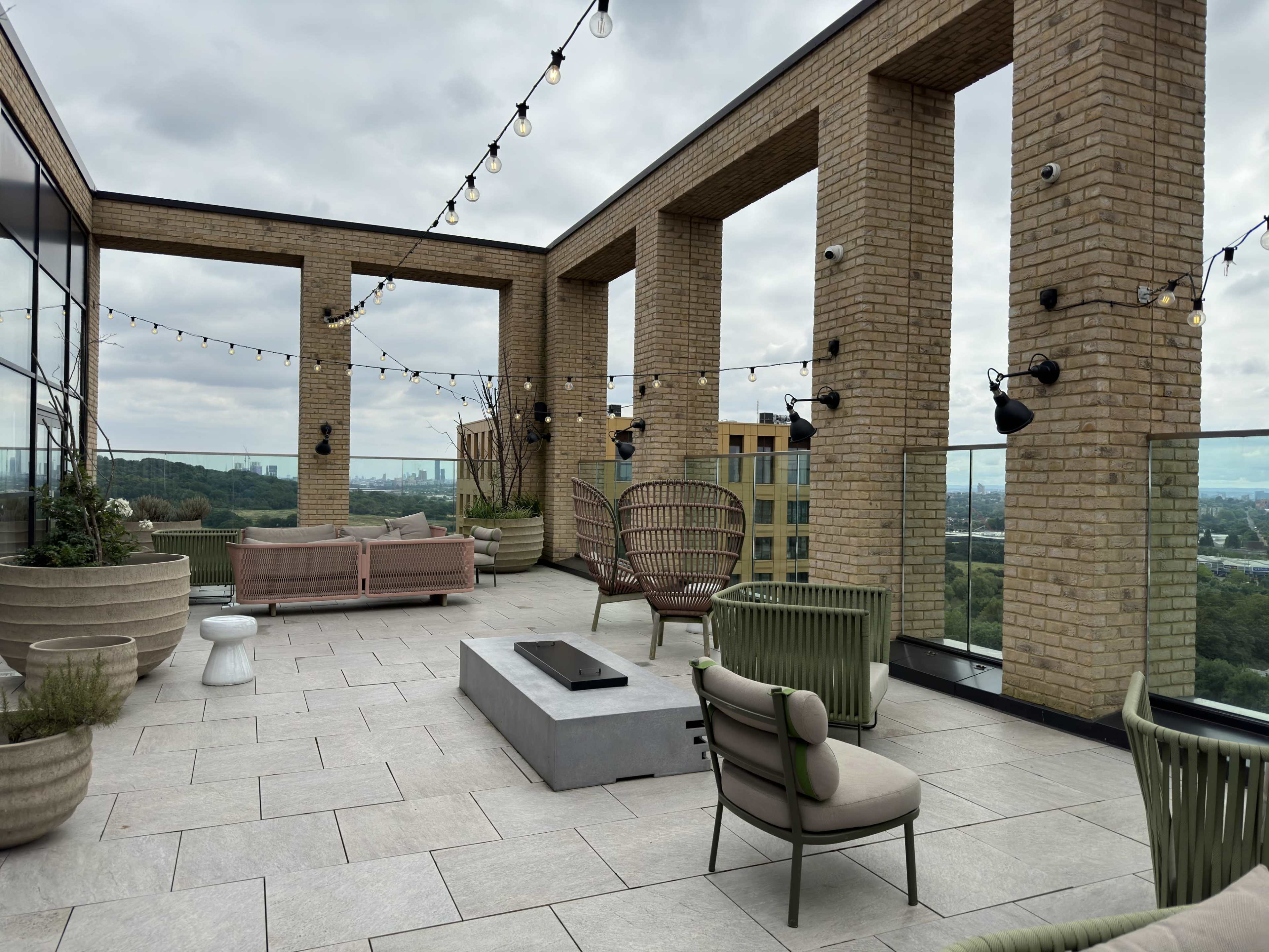 The image shows a rooftop terrace with various seating arrangements and decorative lighting, overlooking a green landscape.