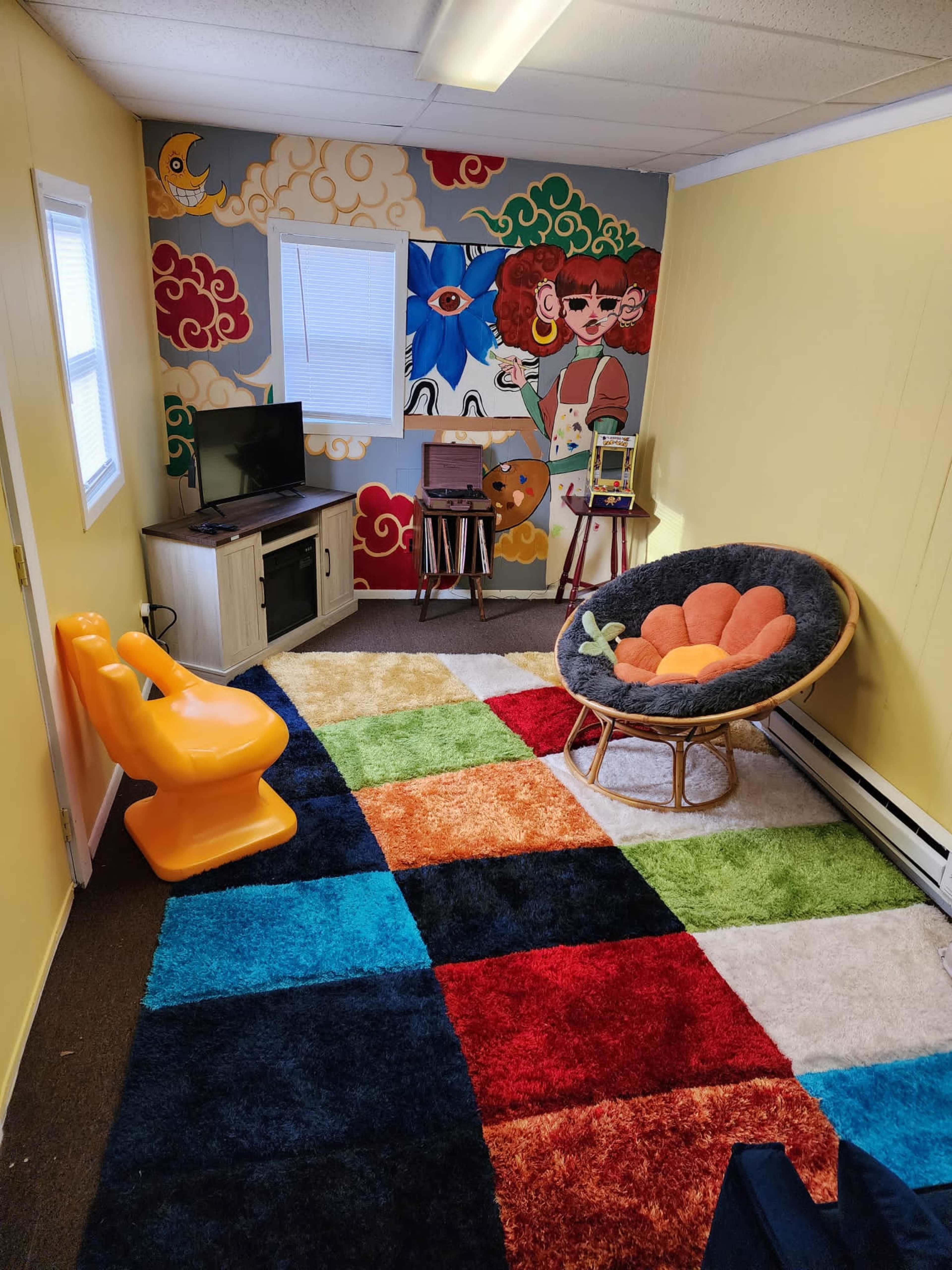 The room features a colorful patterned carpet, a wall mural with cartoon characters, a TV on a cabinet, and a cozy chair.