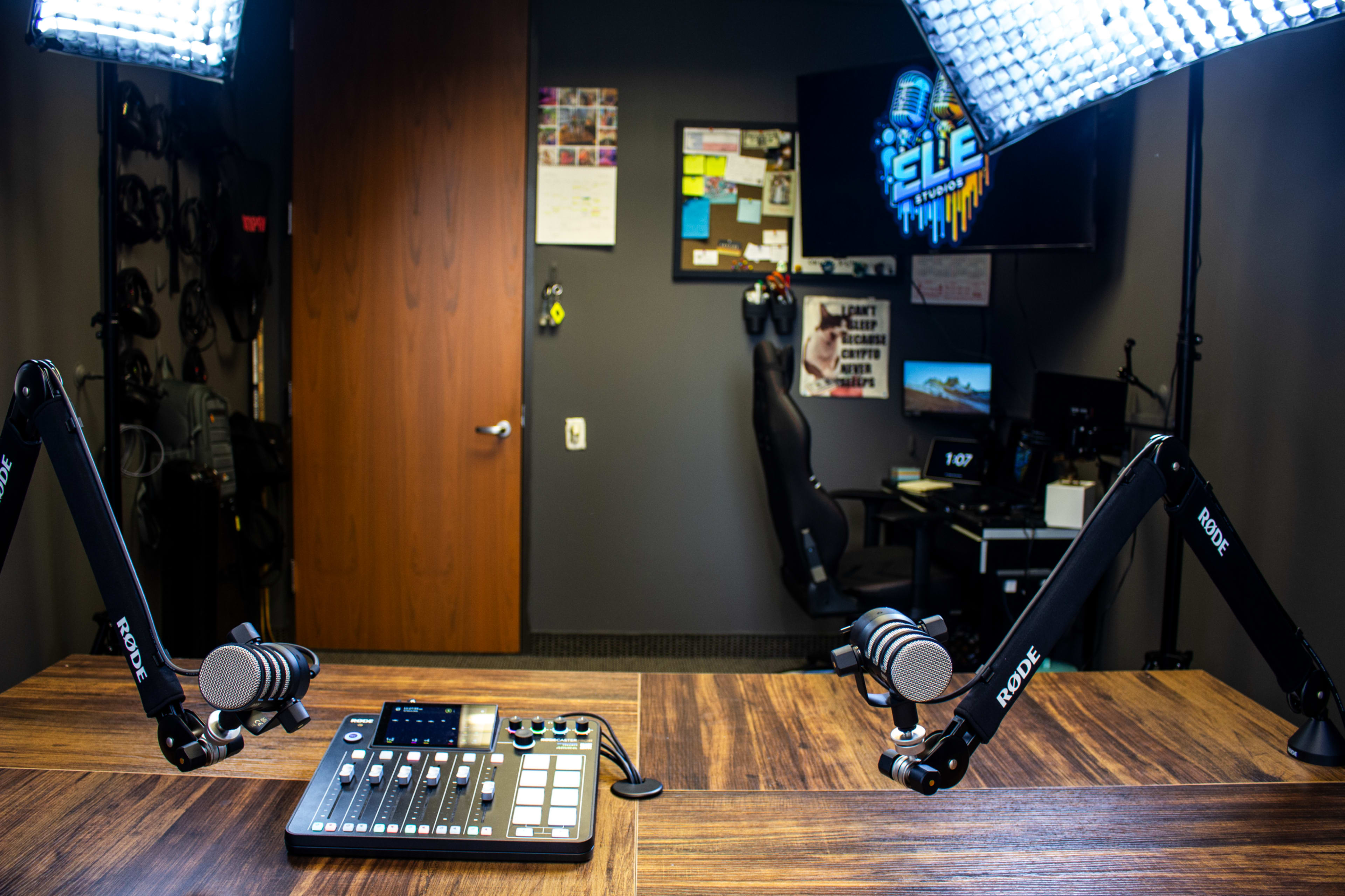 The image shows a broadcasting studio with a wooden table, microphone stands, and a monitor displaying a logo on the wall.