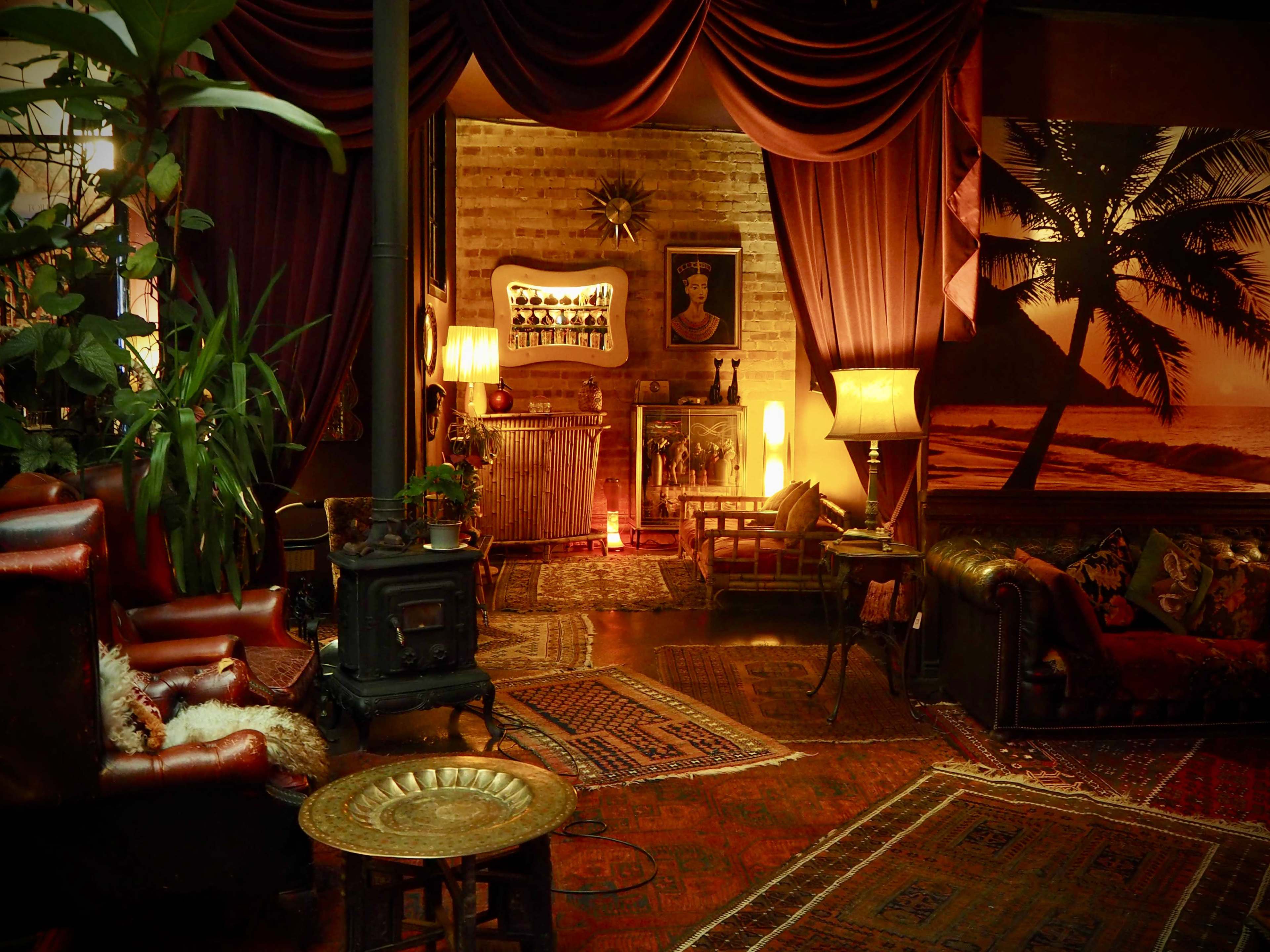 A warmly lit, cozy interior with vintage furniture, plants, and rich textiles, featuring a fireplace and decorative artwork.