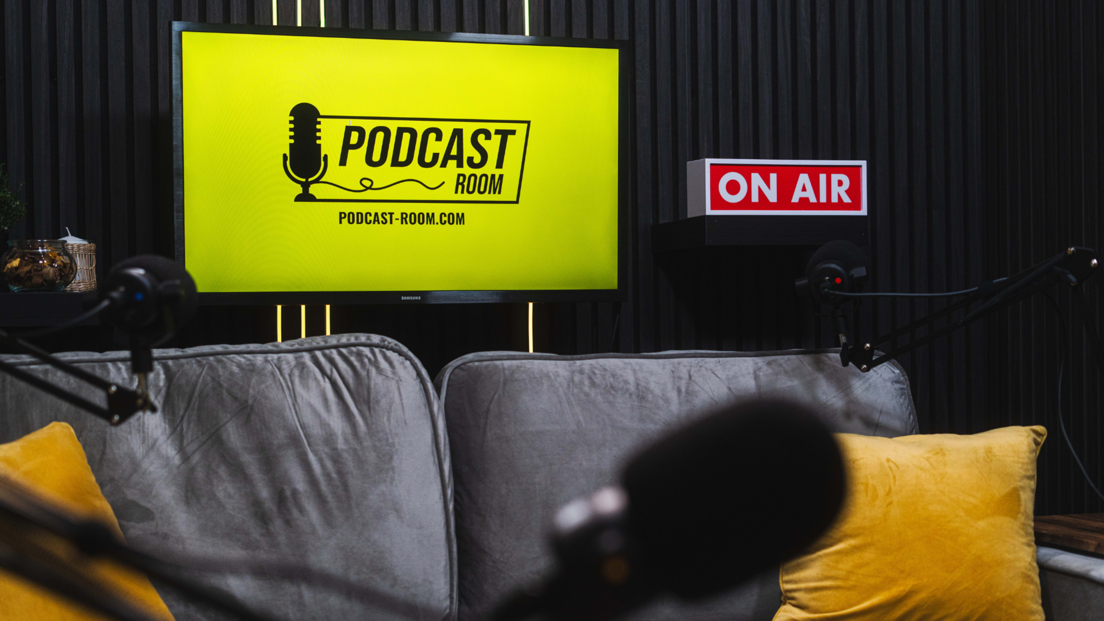 A television screen displaying a podcast logo and a "ON AIR" sign is set against a dark wooden backdrop, with microphones and a sofa in the foreground.