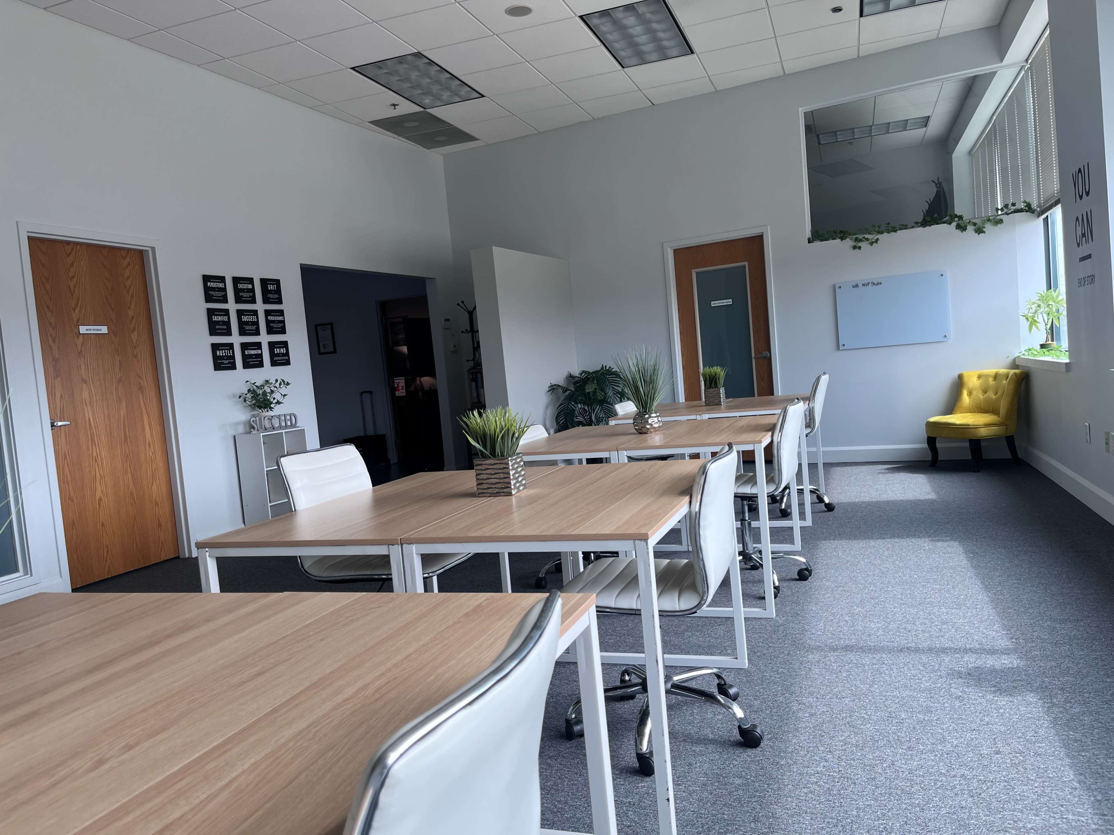 A spacious, well-lit meeting room with several wooden tables and white chairs, along with a yellow armchair in the corner.