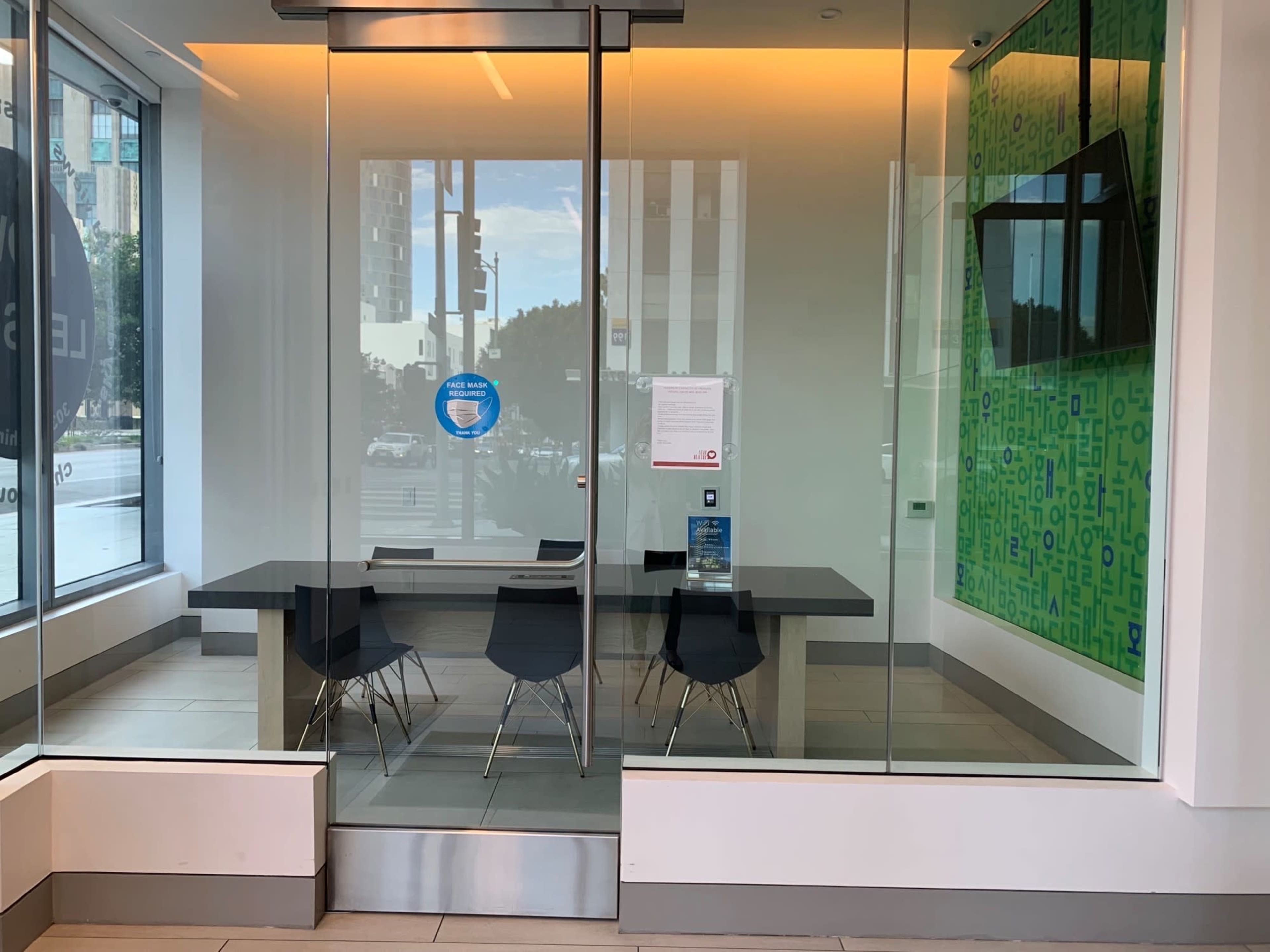The image shows a glass-walled meeting room featuring a black table and four black chairs, with a television mounted on the wall and a view of a city street outside.