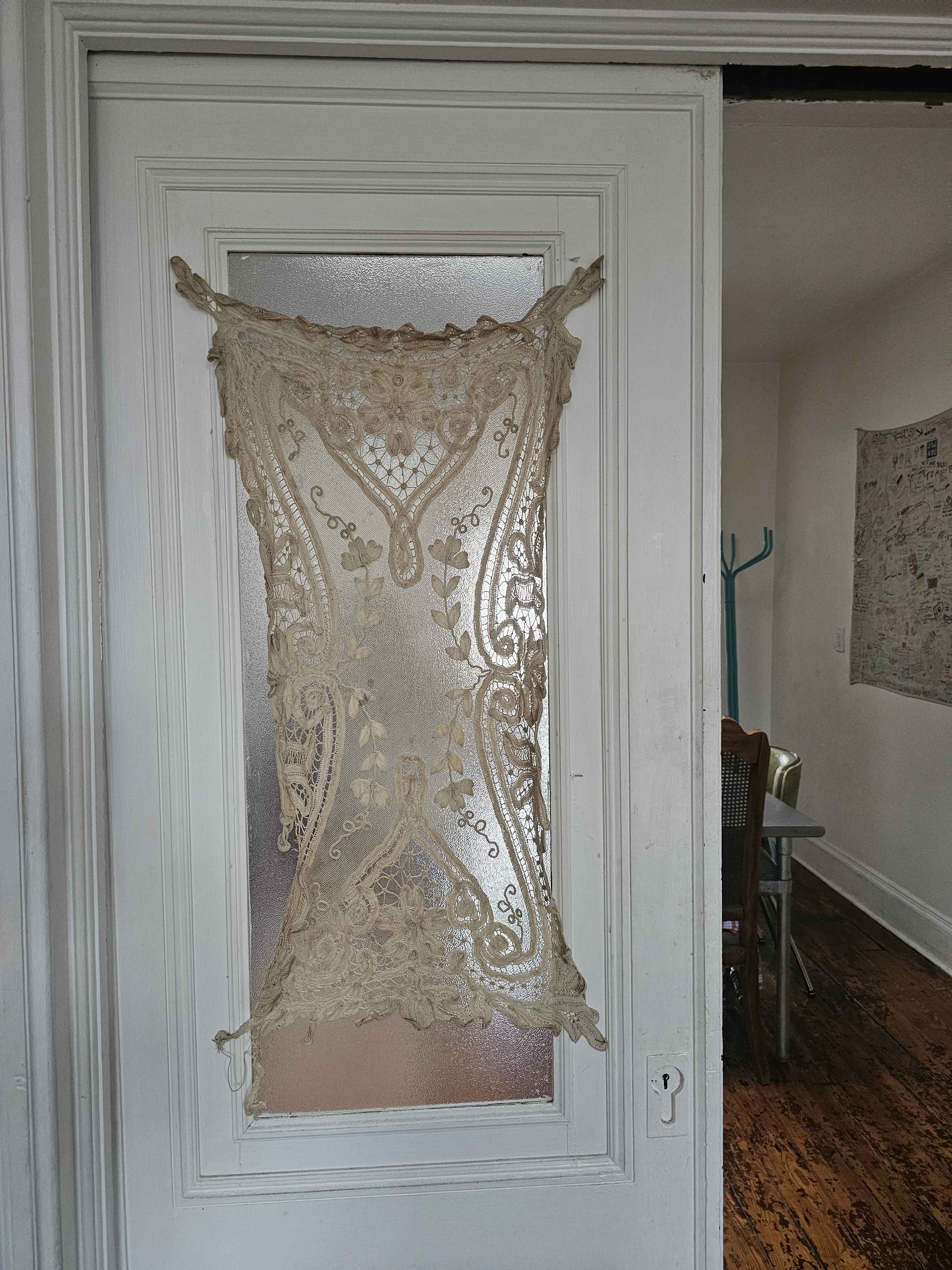 A lace curtain is hung on a frosted glass door in a room with wooden flooring and visible furniture in the background.