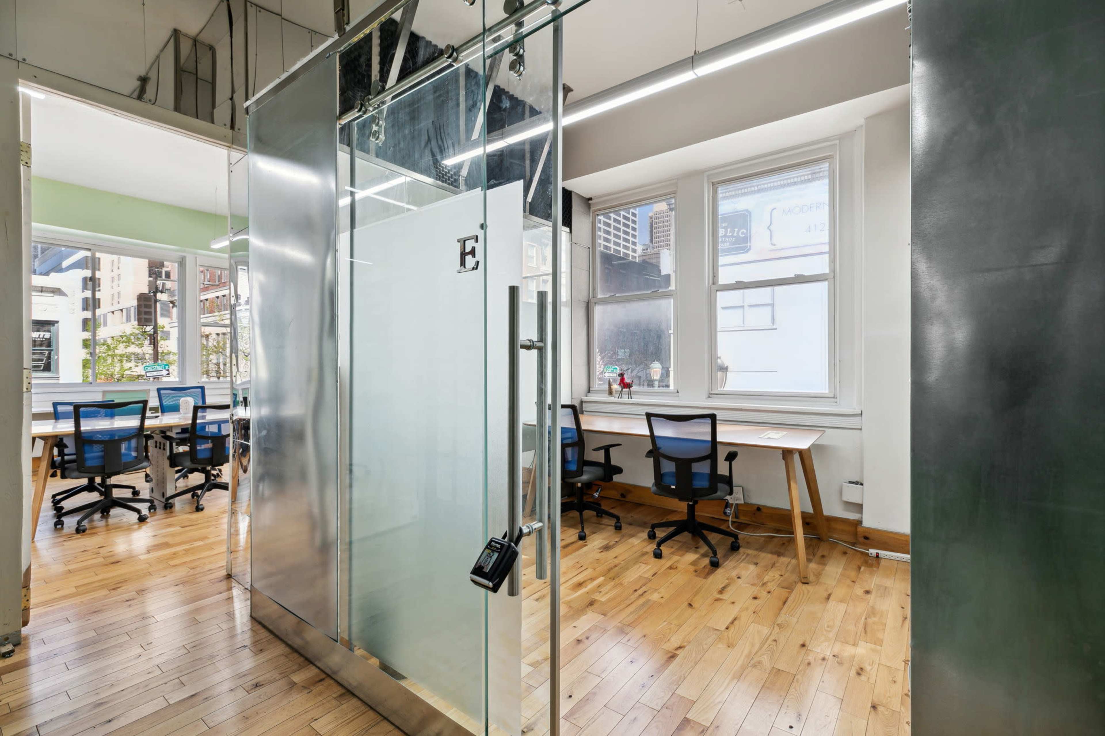 An office space featuring a glass-walled conference room adjacent to workstations with wooden flooring and large windows.