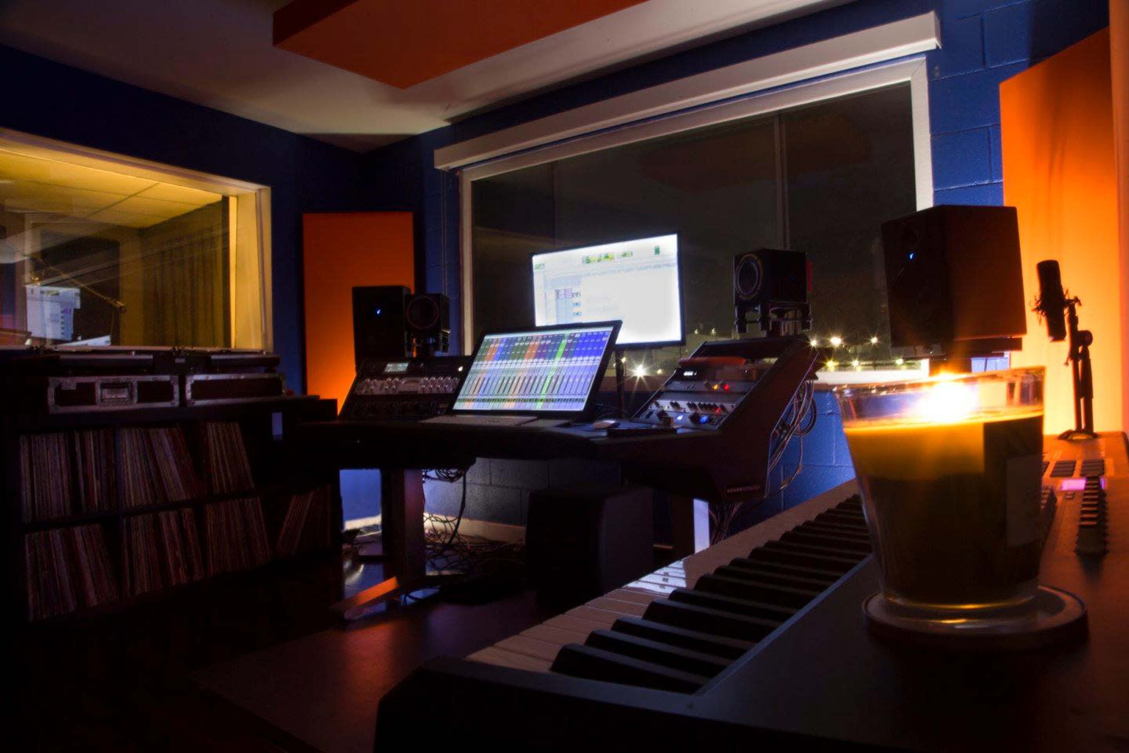 A recording studio features a computer workstation with digital audio software, speakers, and a collection of vinyl records, illuminated by ambient lighting and a candle on a desk.