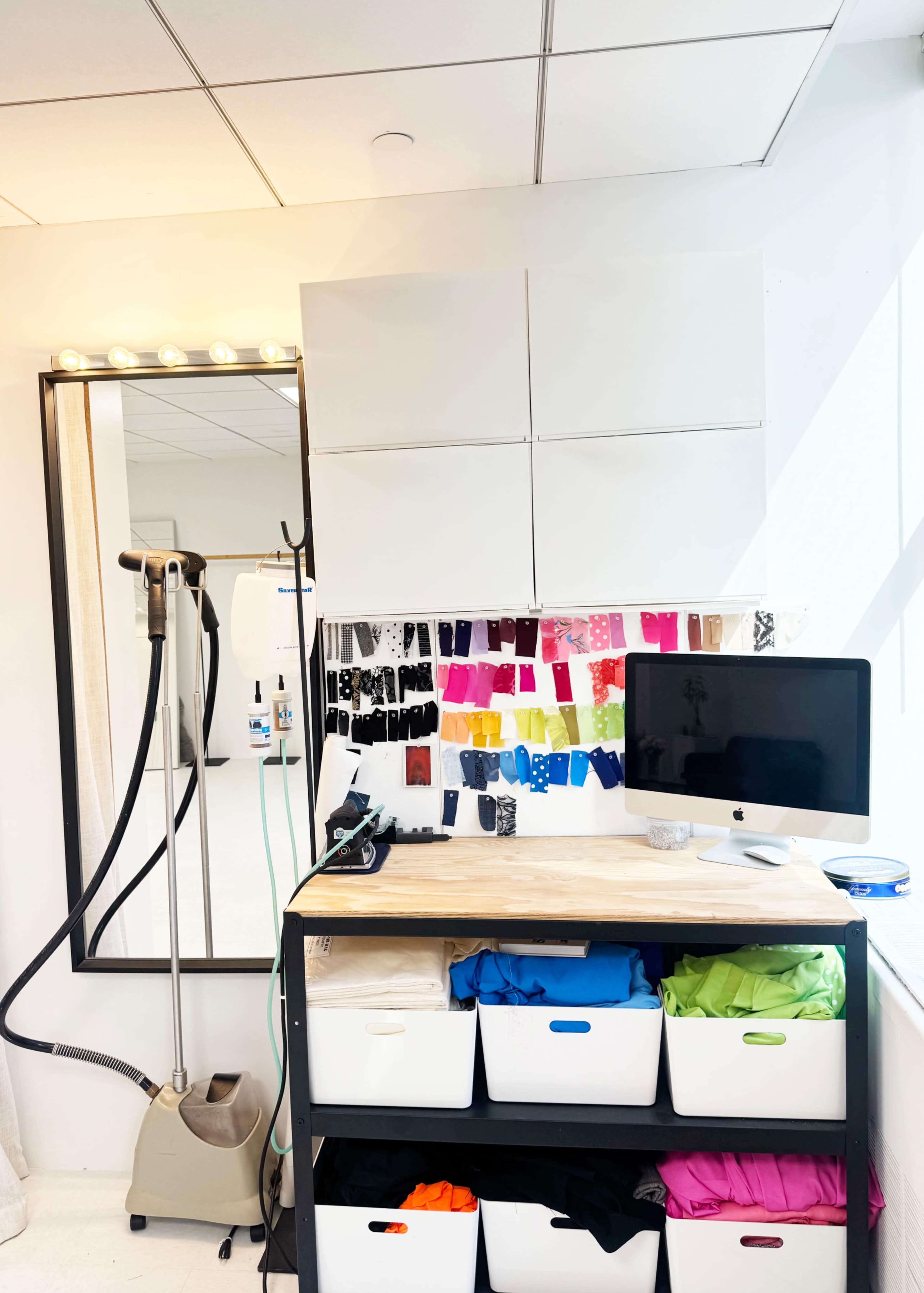 The image shows a well-organized workspace featuring a mirror with lights, a steaming machine, colorful fabric swatches displayed on the wall, and storage bins filled with various colored items beneath a wooden tabletop.