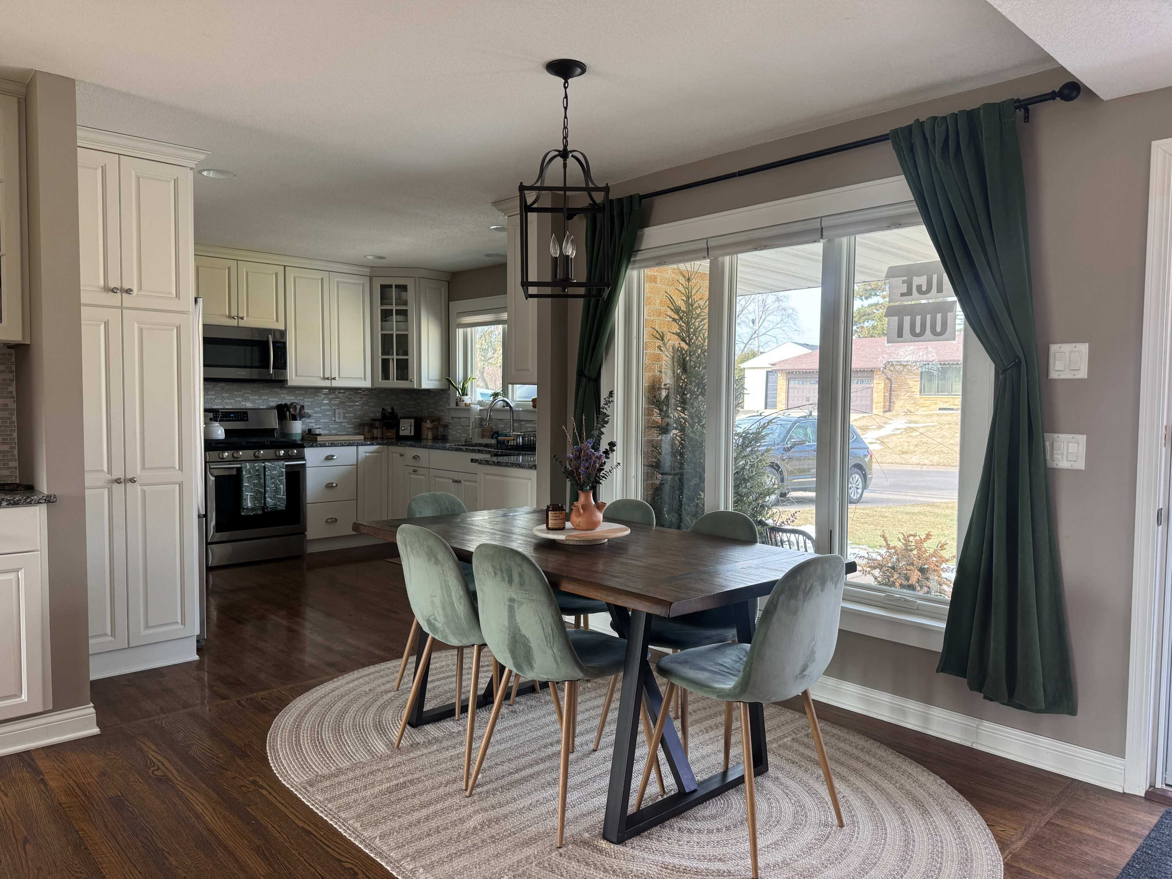 The image shows a modern dining area with a wooden table and green chairs, adjacent to a kitchen with white cabinets and a large window facing a front yard.