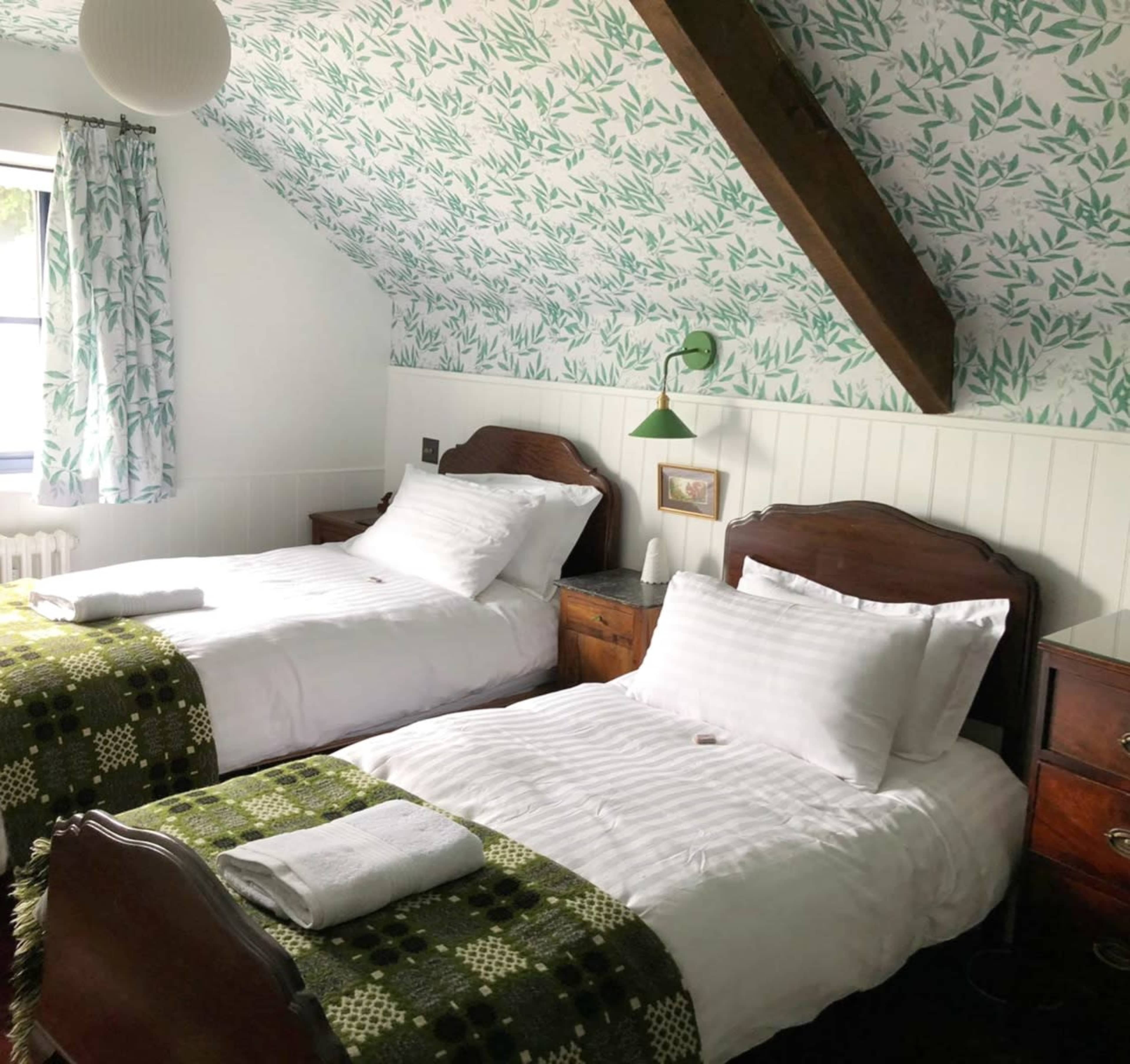 The room features two twin beds with white linens and green-patterned throws, flanked by wooden nightstands under a sloped ceiling adorned with leafy wallpaper.