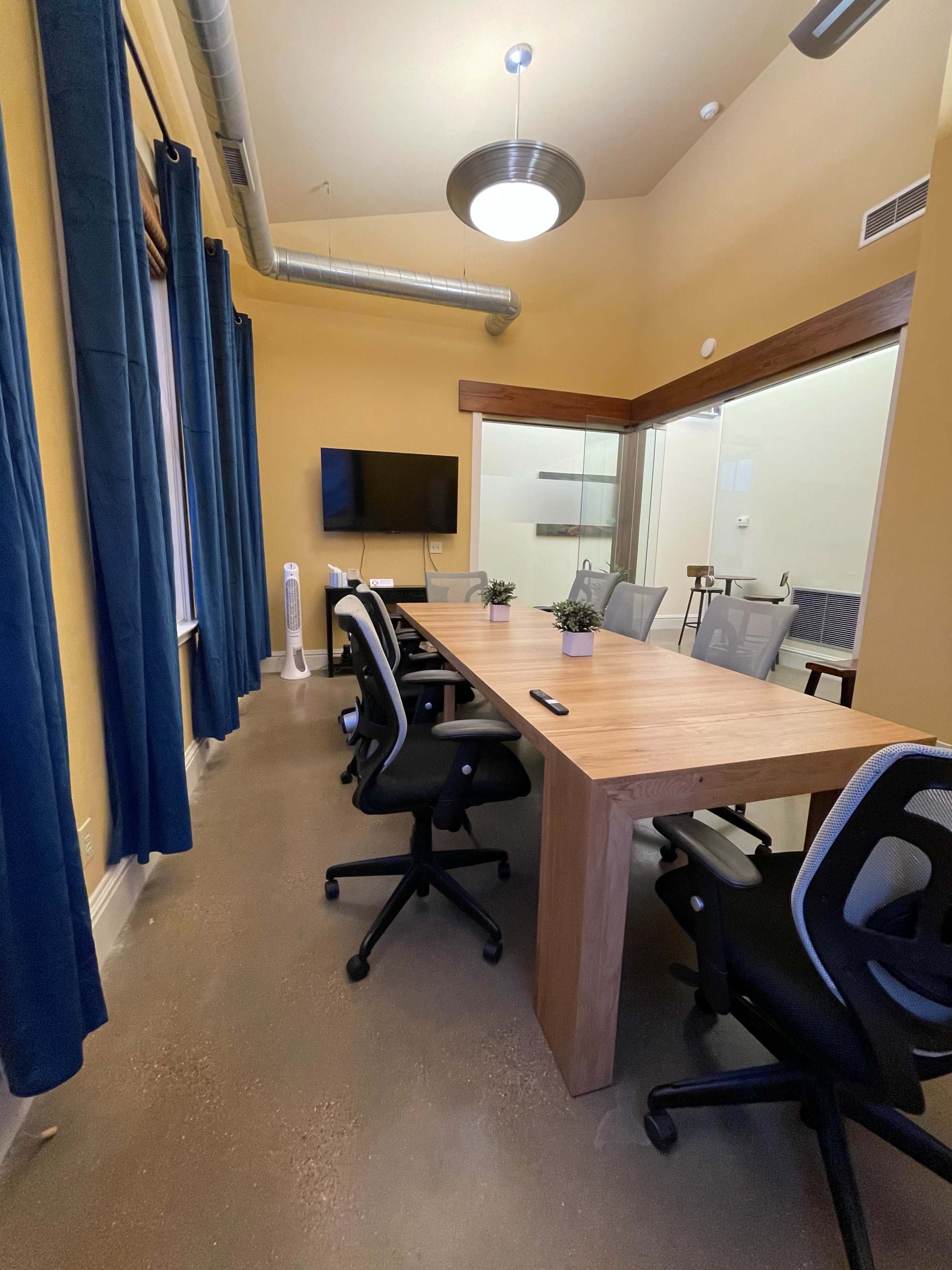 The image shows a modern conference room with a wooden table, black office chairs, a television mounted on the wall, and large windows covered by blue curtains.