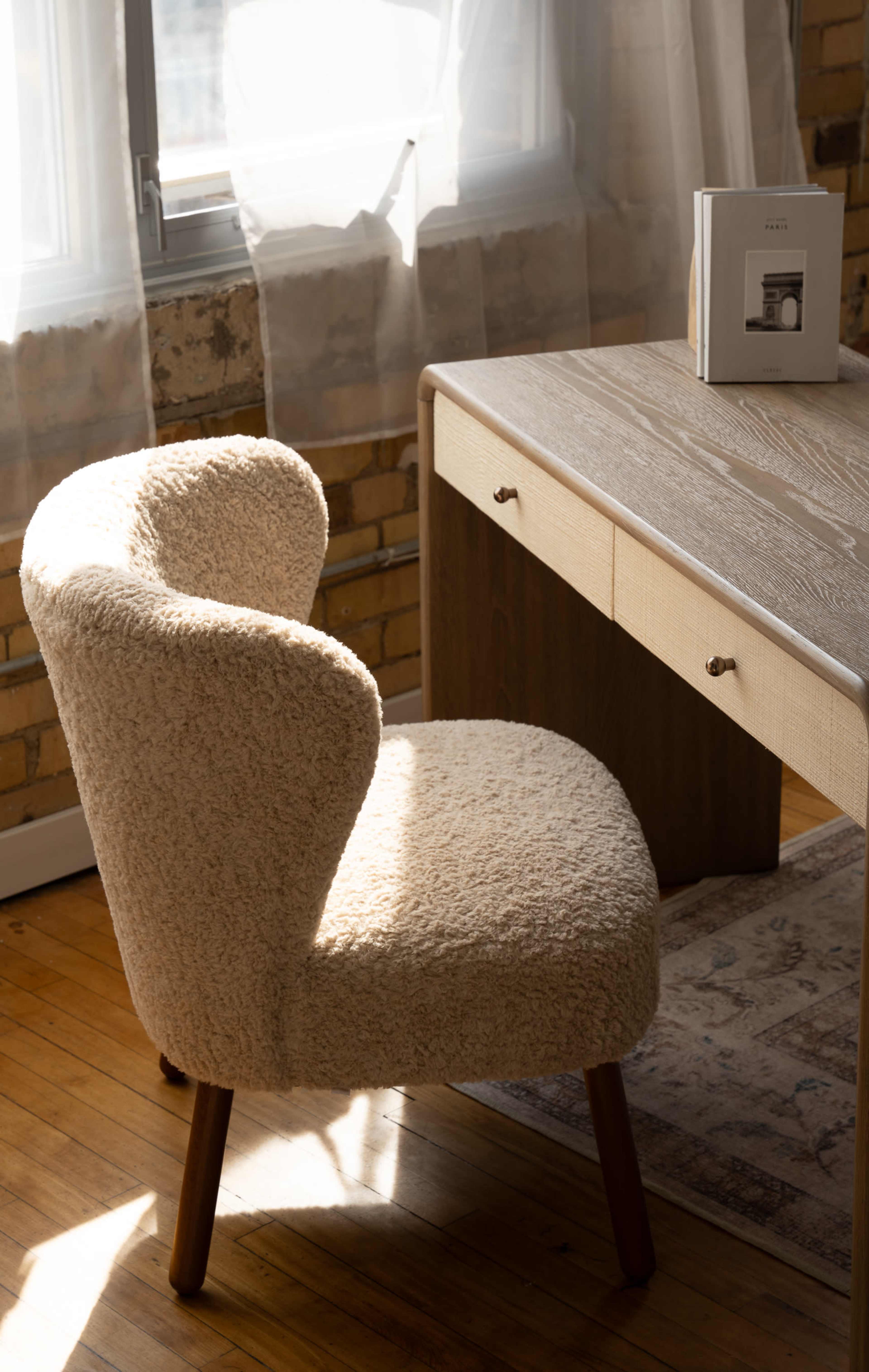 A textured, cream-colored chair sits beside a wooden desk in a brightly lit room with large windows.