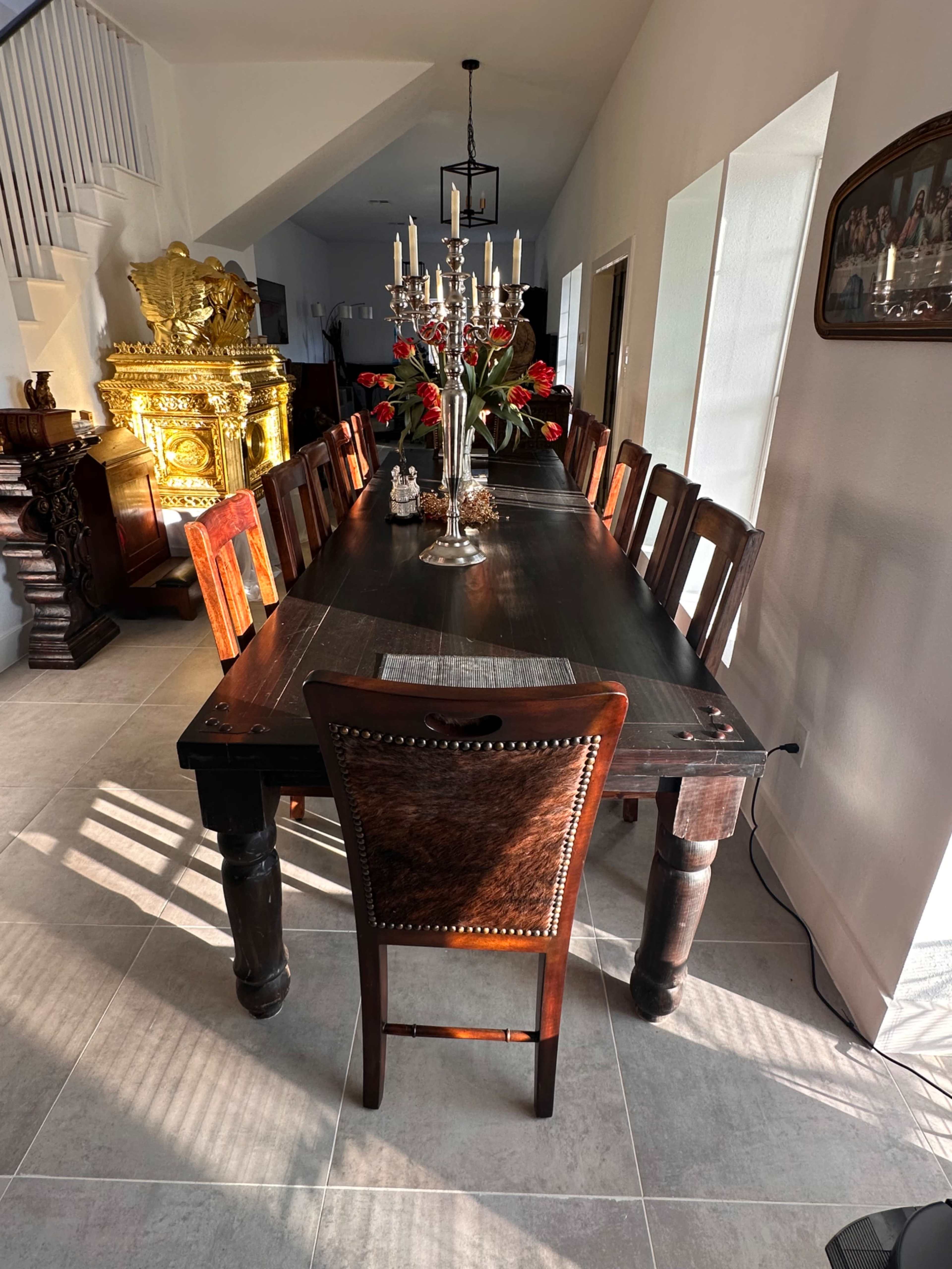 A long wooden dining table with multiple chairs is adorned with a vase of red roses and a candelabra, set in a bright room with white walls.