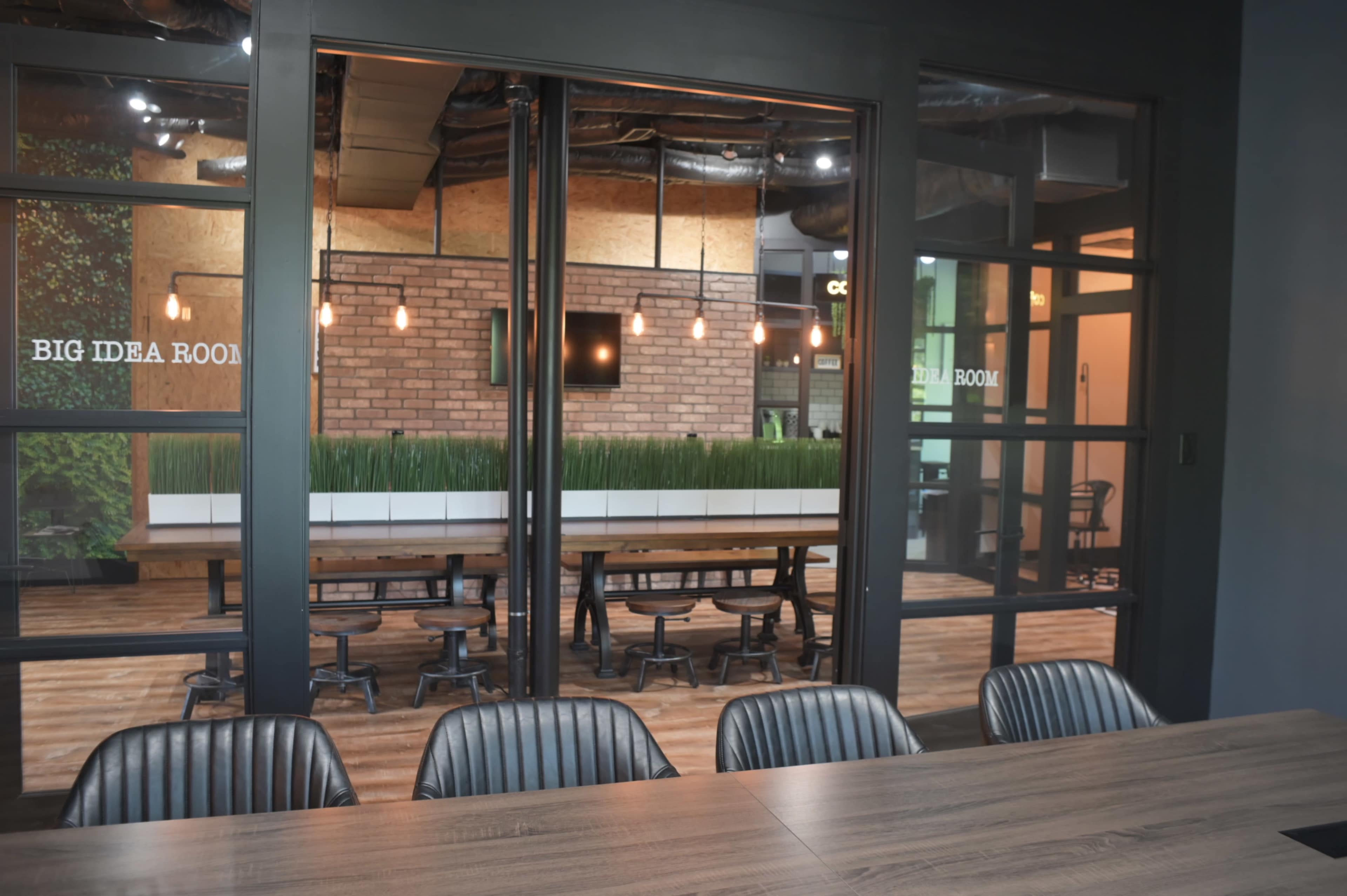 A glass-walled conference room with a large wooden table, overlooking a spacious meeting area decorated with greenery and pendant lights.