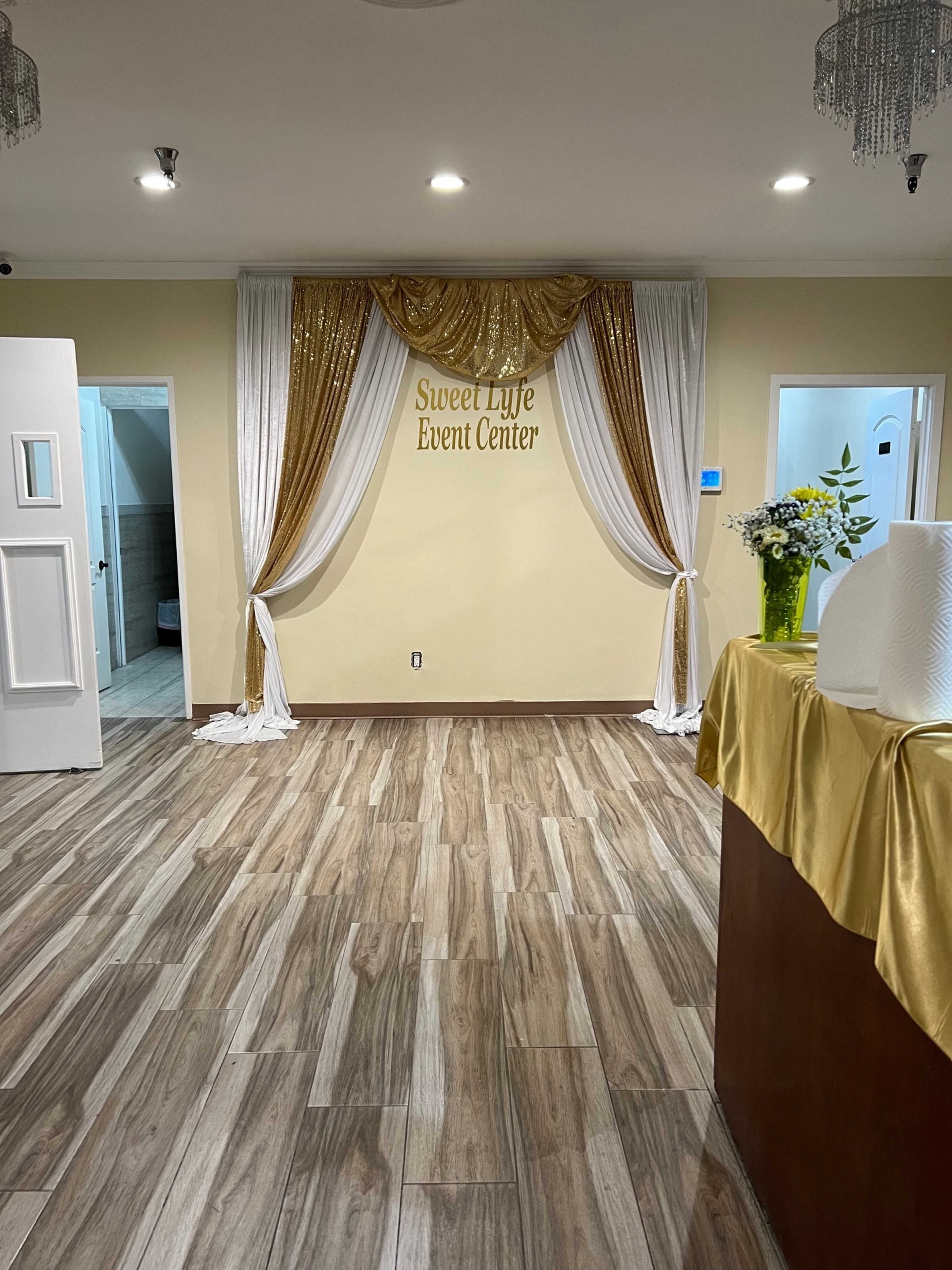 The interior of the Sweet Life Event Center features a decorative backdrop with gold and white drapes, a floral arrangement, and wooden flooring.