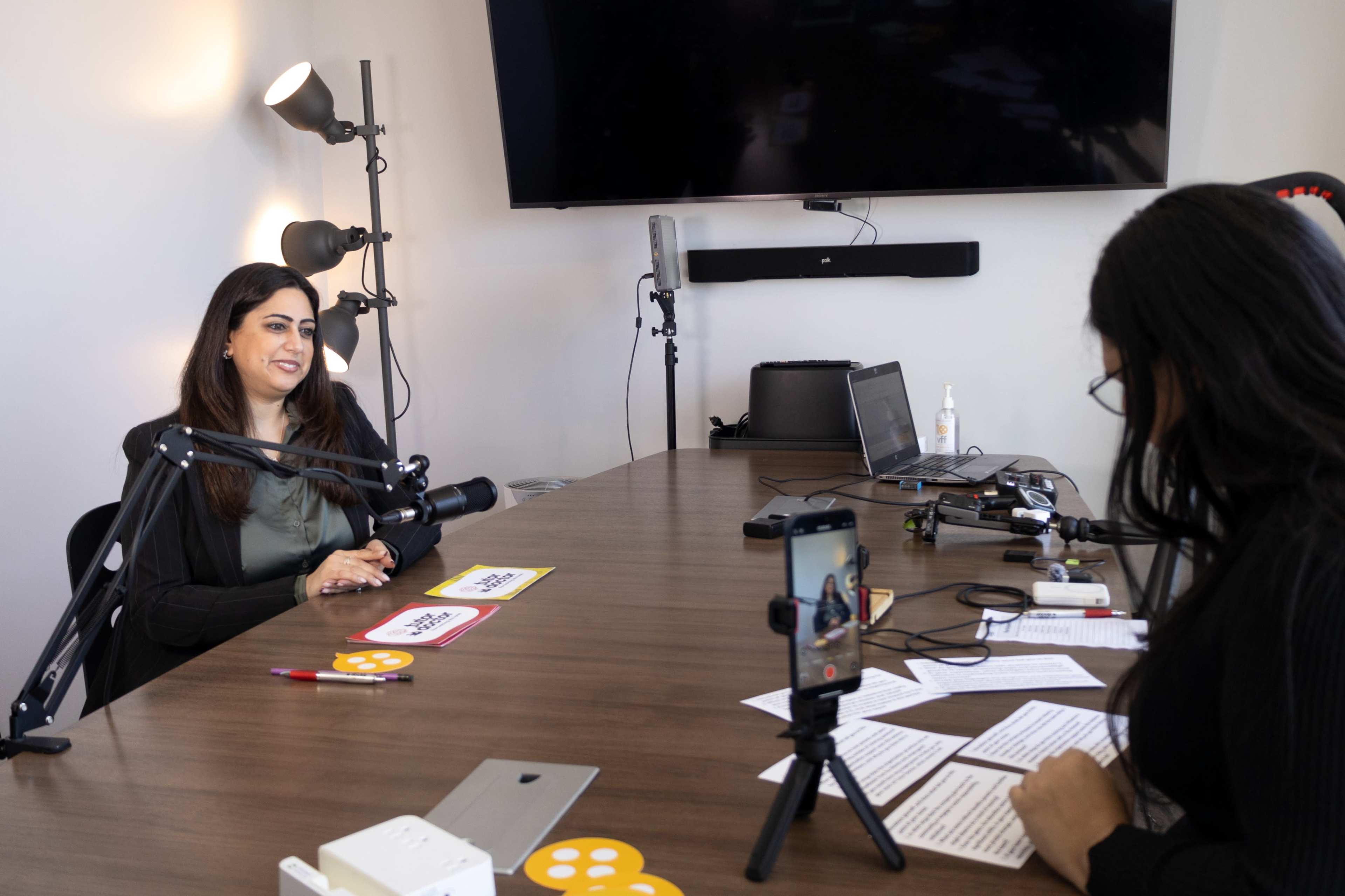A woman sits at a table with a microphone in front of her while another woman records her with a smartphone in a modern office setting.