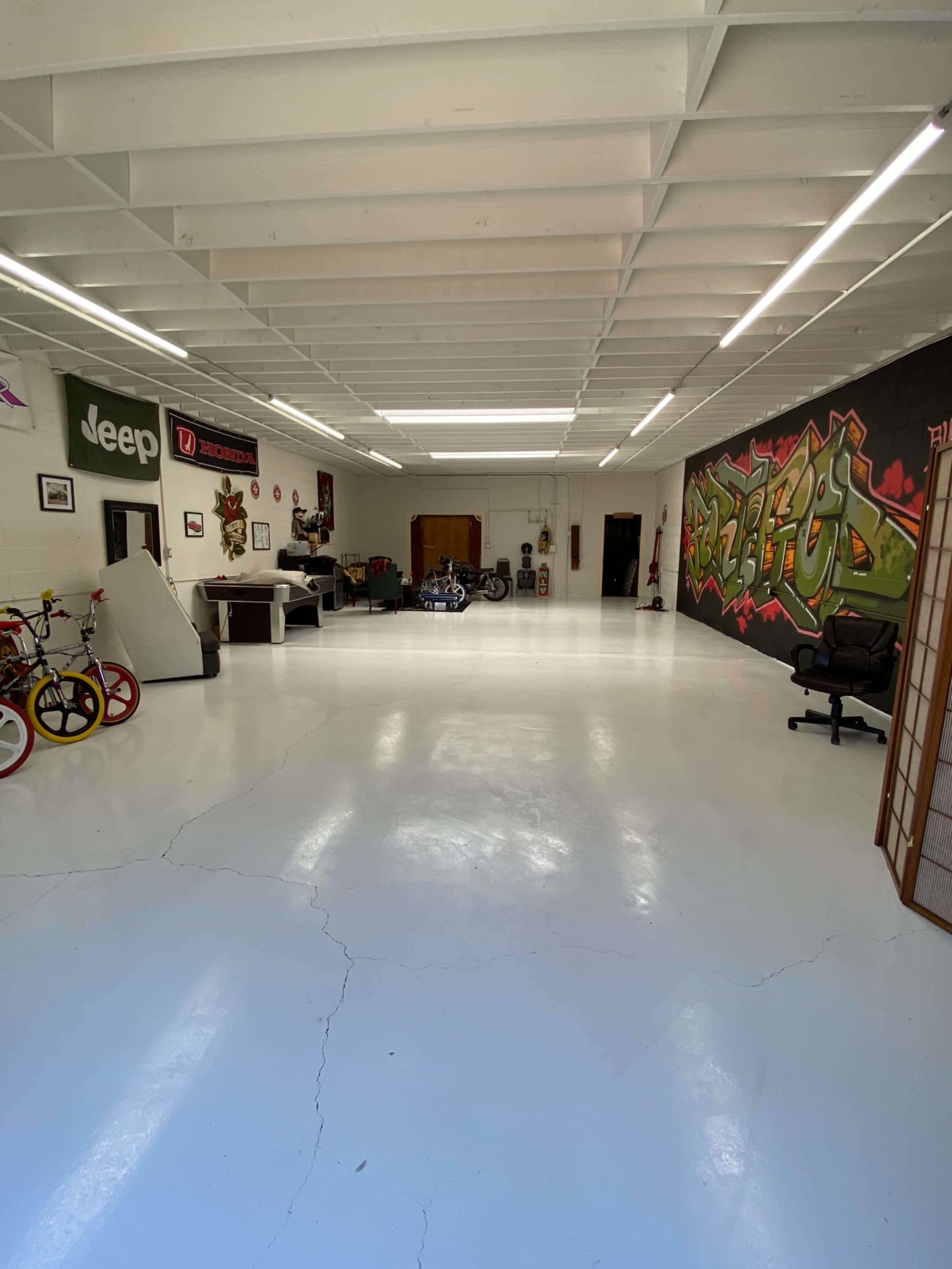 The image shows a spacious garage or workshop with a polished white floor, featuring bikes, motorcycles, and graffiti art on the walls.