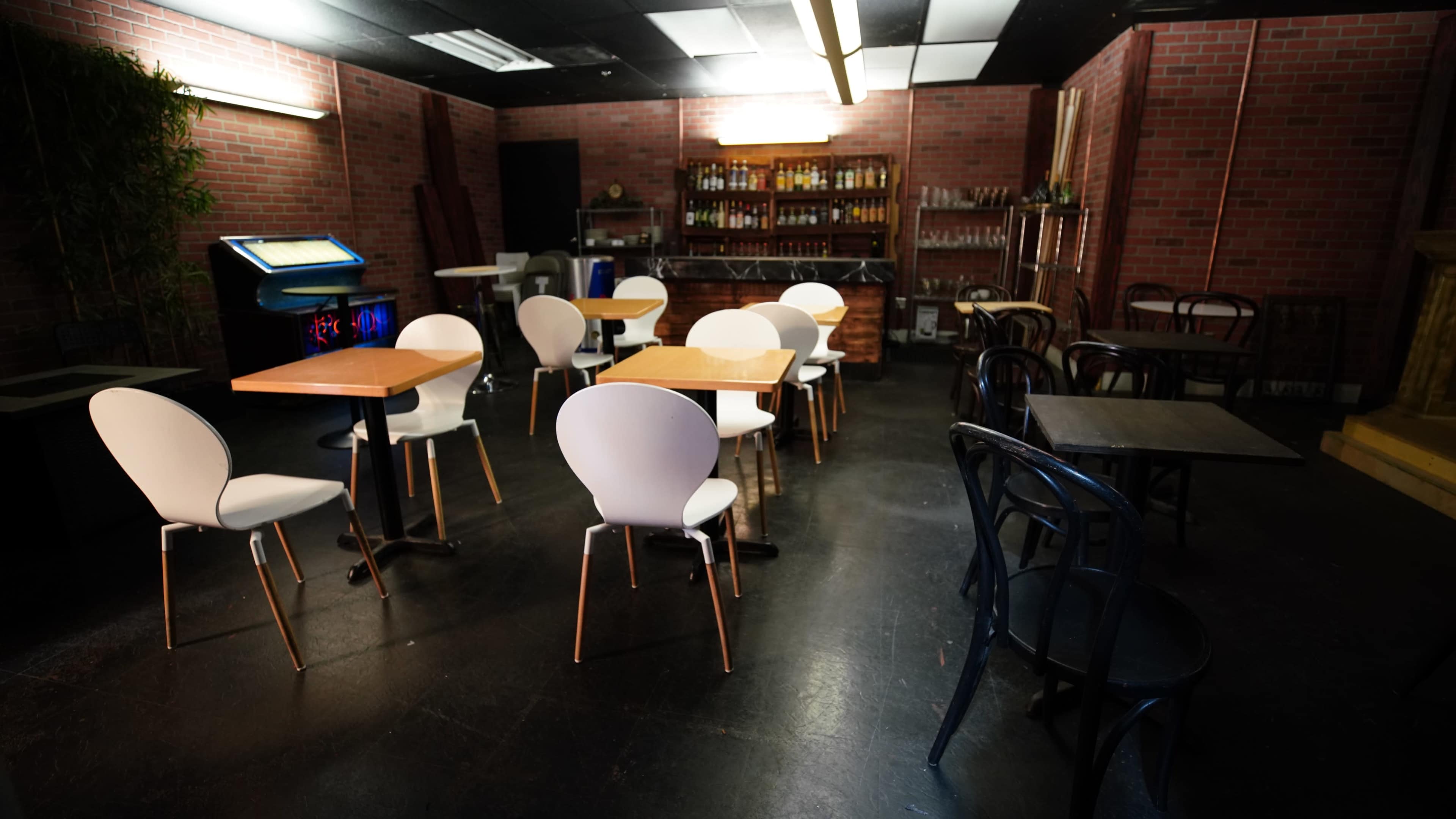 An empty bar area with wooden tables and a mix of white and black chairs, complemented by a brick wall and a shelf filled with bottles in the background.