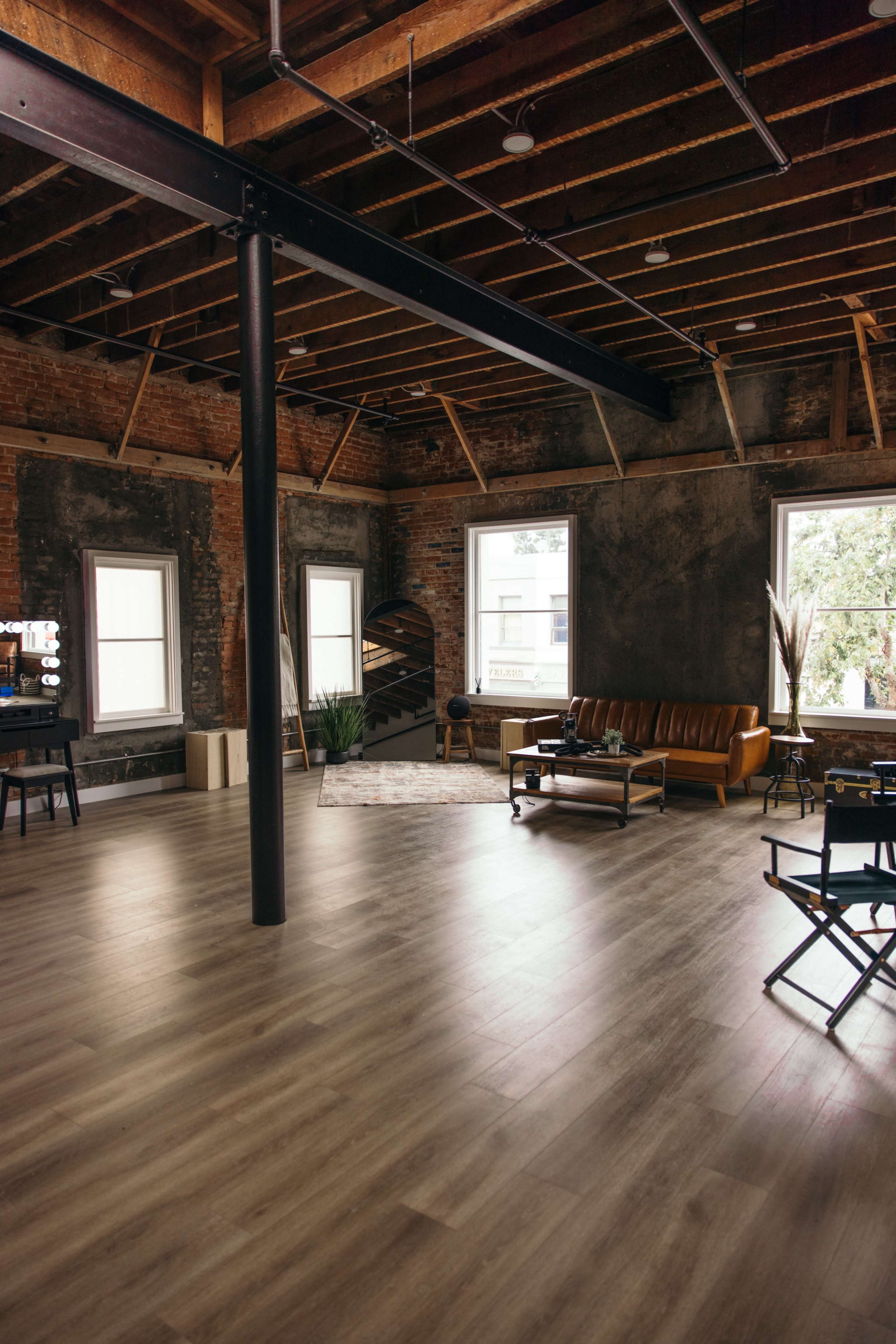 A spacious room with exposed brick walls, wooden beams, and large windows, featuring a leather sofa, a staircase, and a few plants.
