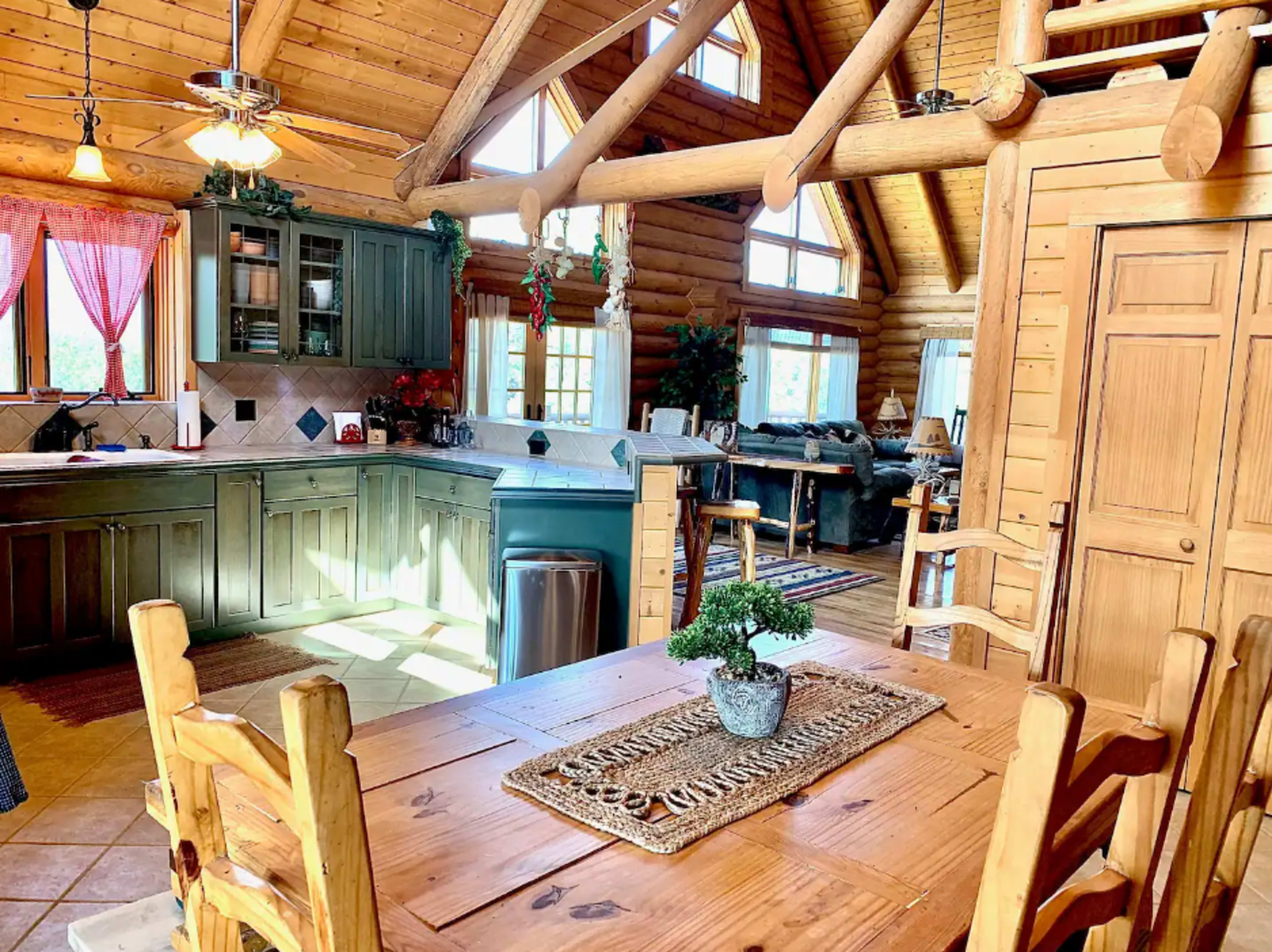 The image shows a wooden kitchen and dining area in a log cabin, featuring a table, chairs, and large windows allowing natural light.