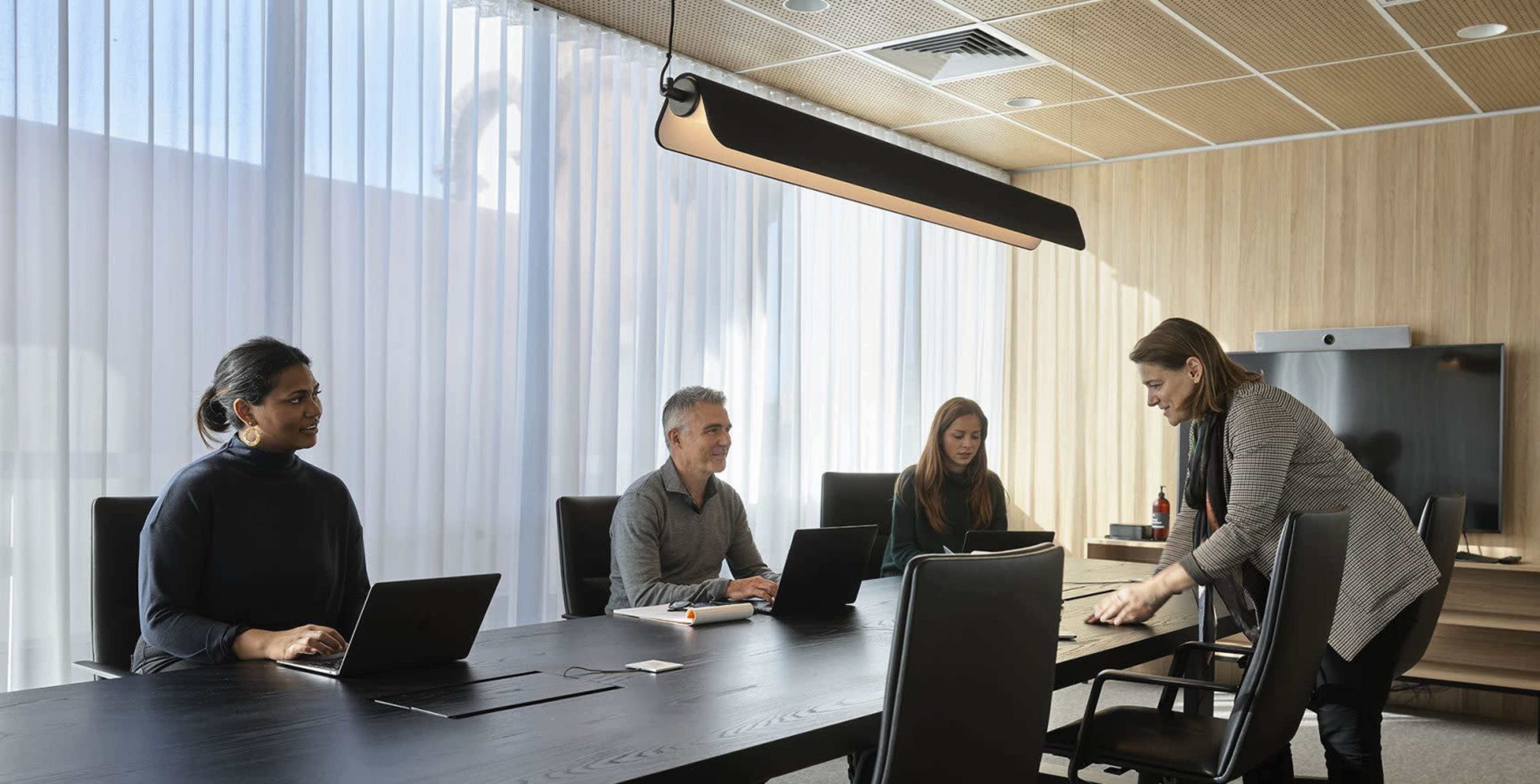 A group of four professionals is seated around a conference table in a well-lit meeting room, with one person standing and arranging materials.