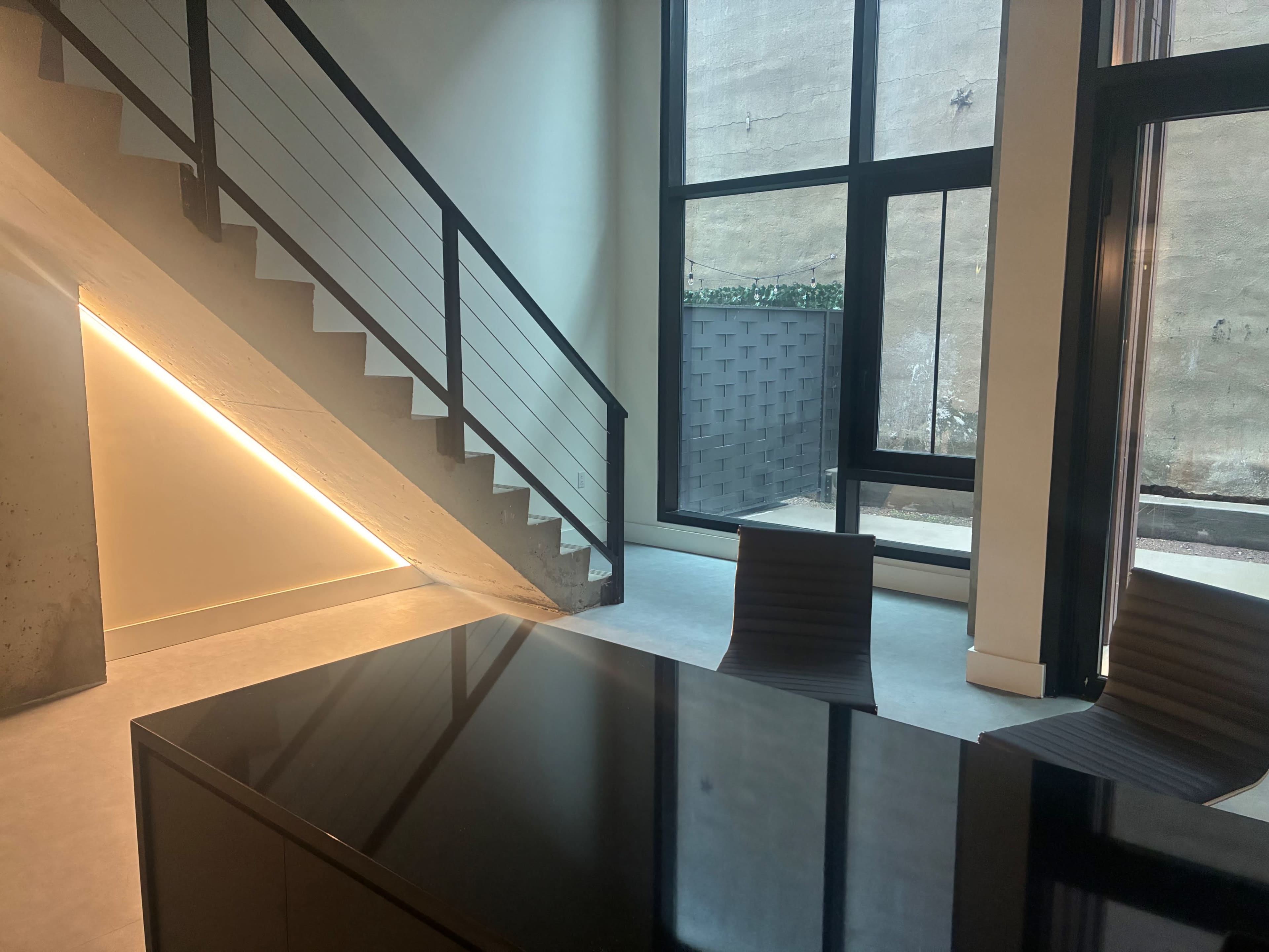 The image shows a modern interior office space featuring a staircase, a large black table, and glass doors leading to an outdoor area.