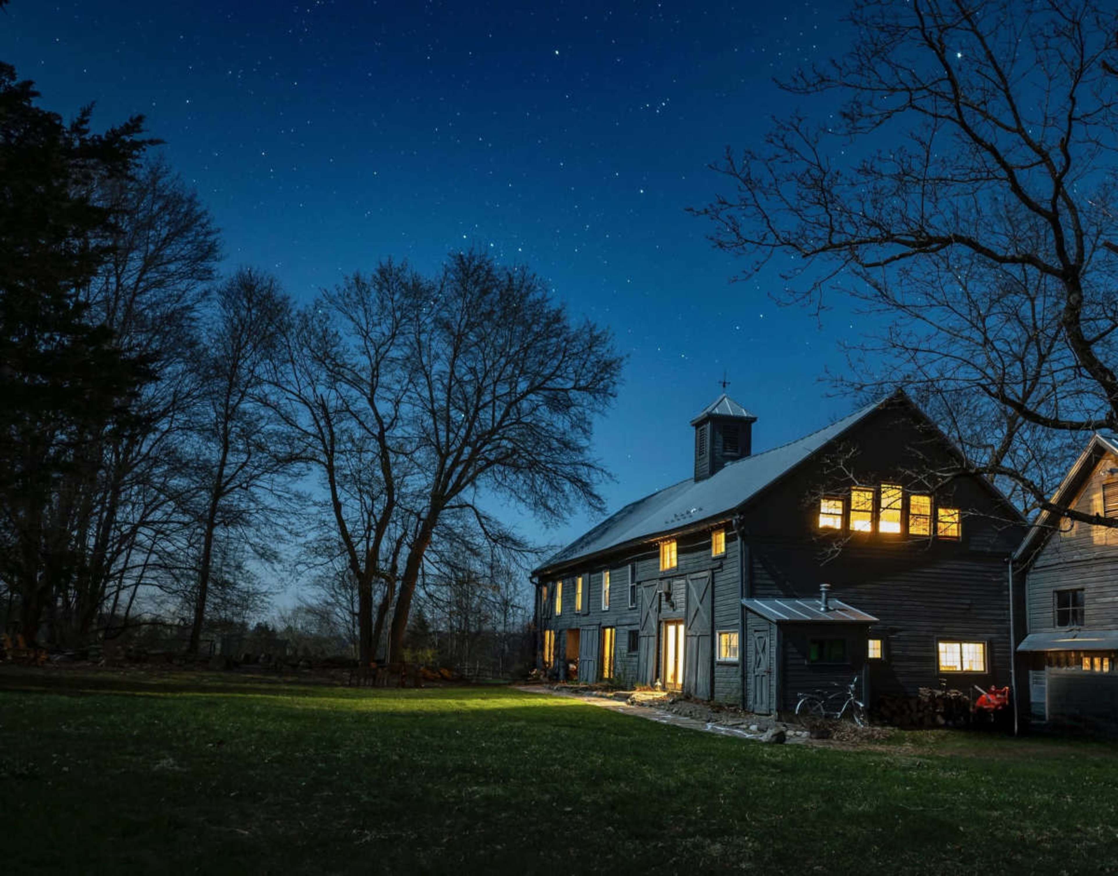 A dark wooden barn stands under a starry night sky, illuminated by warm lights from its windows and doors.