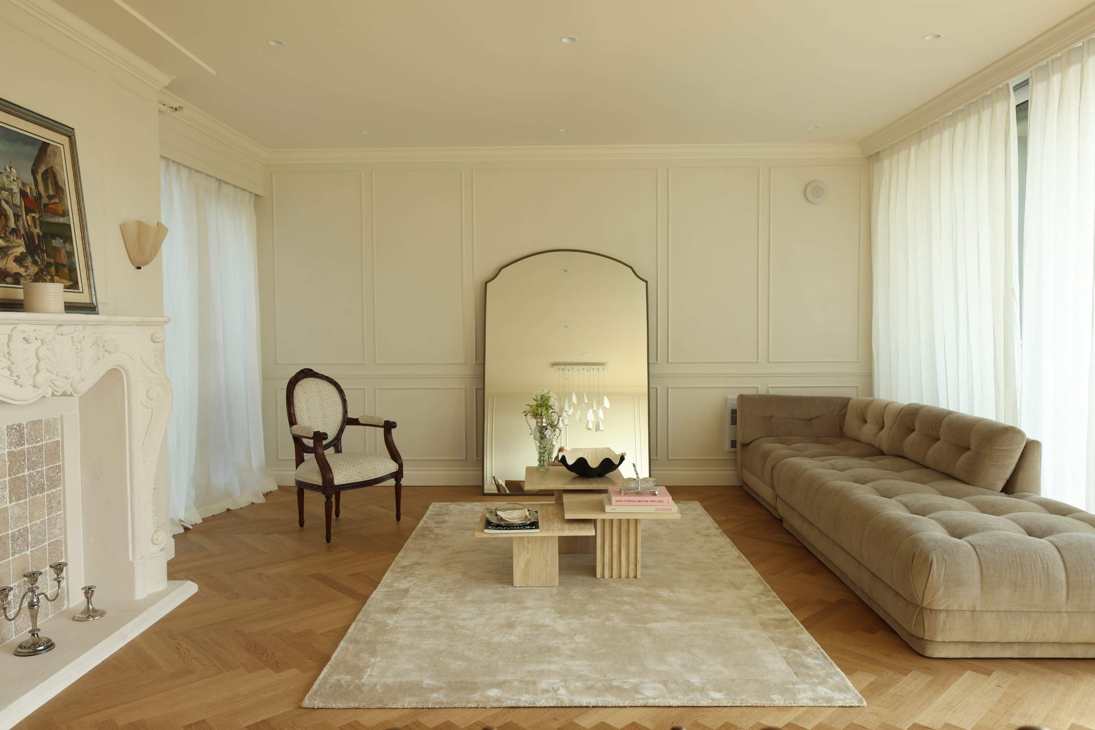 A modern living room features a beige sofa, a coffee table made of stacked wooden cubes, and a large mirror, with hardwood flooring and soft natural light filtering through sheer curtains.