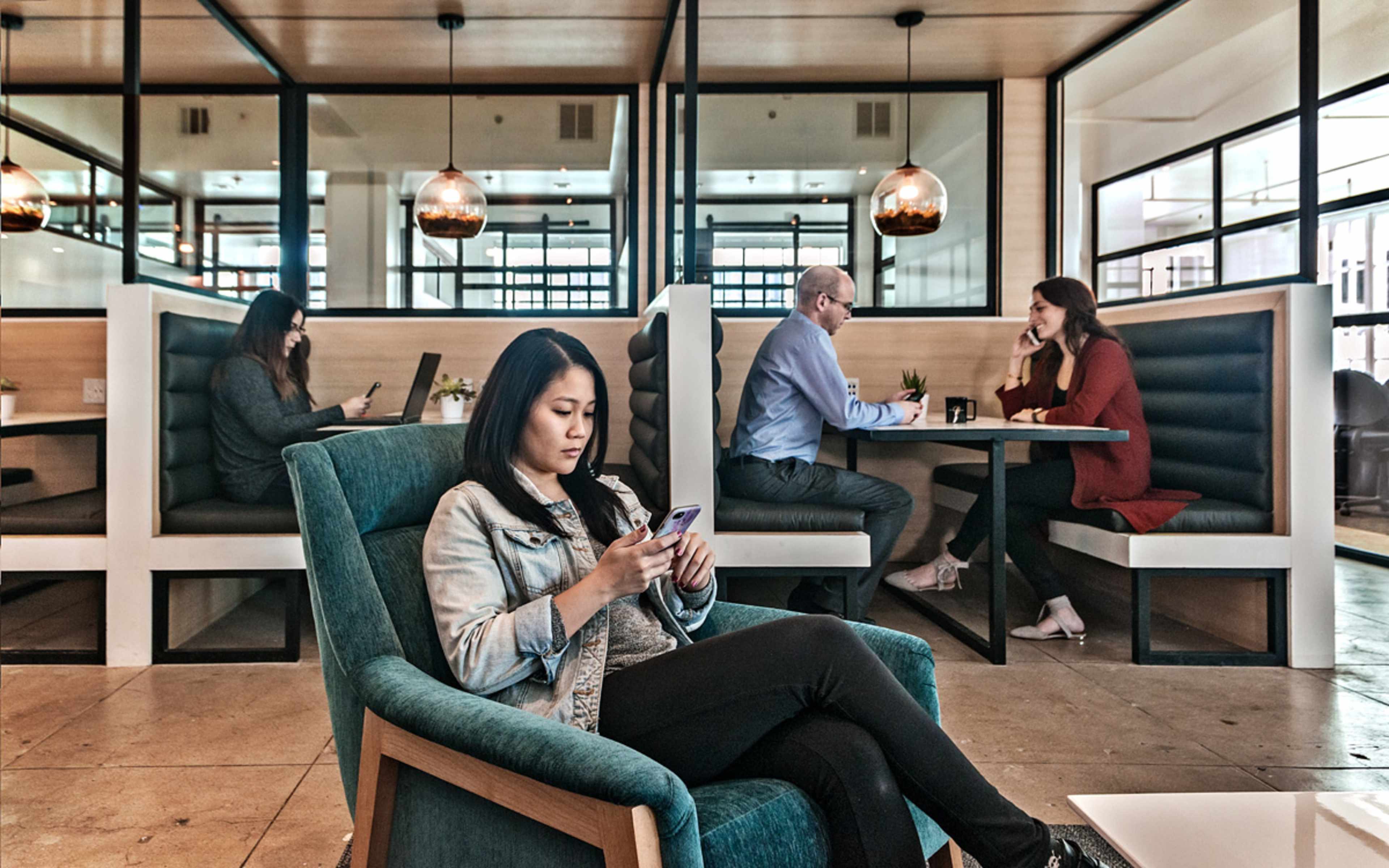 A woman sits in a teal chair using her phone while two people engage in conversation at a nearby table, and another person works on a laptop in the background.